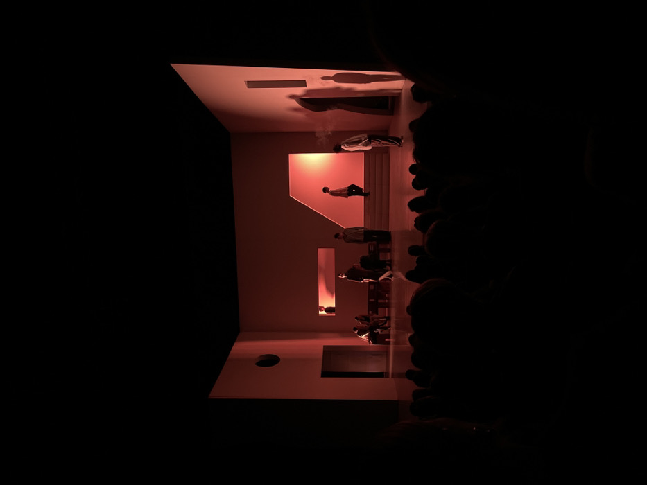 A theatrical stage performance is depicted within a theatre in Paris, France. The scene focuses on a minimalist stage set composed of light-colored, angular walls under intense red and orange illumination, creating stark contrasts and pronounced shadows.
On the stage, seven human figures are visible. Stage left, two individuals are seated on a low bench while a third stands nearby. Towards the center, two additional figures stand, one appearing to wear dark clothing. Further right, one individual ascends a prominent staircase structure with their back to the viewer. Another figure stands near the far right wall, facing right, exhaling a visible plume of smoke or vapor. A large, dark silhouette, possibly an eighth figure or a prop, is visible behind a narrow vertical opening in the right wall, casting a long shadow. The figures are dressed in simple, unadorned clothing. Their postures suggest static composition rather than dynamic interaction.
The stage design includes a large rectangular doorway and a circular cutout on the left wall, a horizontal light aperture on the back wall, and the aforementioned vertical light aperture on the right wall. The foreground of the image shows the silhouetted heads and shoulders of numerous audience members, indicating a full auditorium viewing the performance.