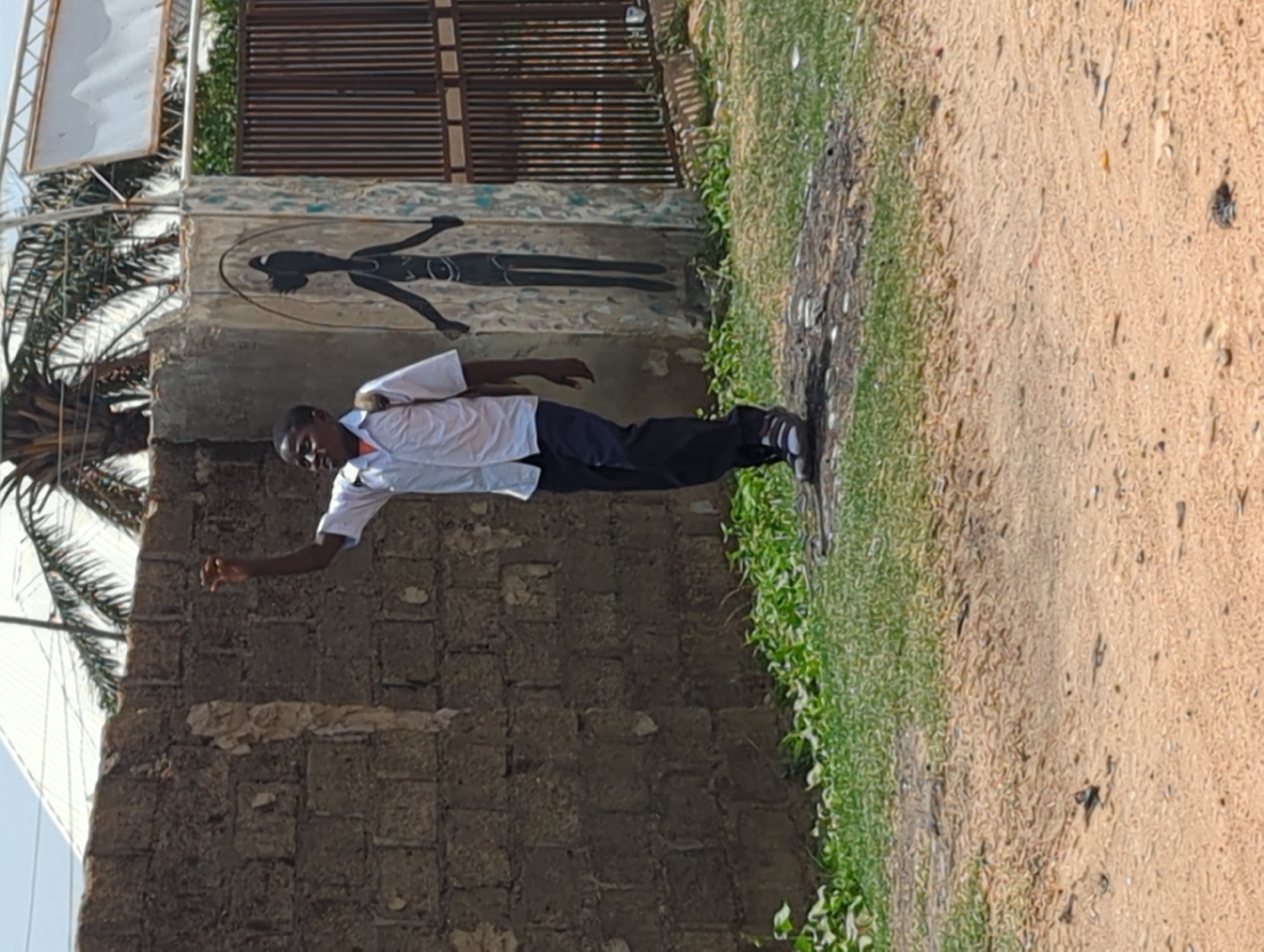 A young man stands in Narayi, Nigeria, in front of a concrete structure that has a silhouette of a person painted on it. He is wearing a white shirt, black pants, and sneakers. The ground is dirt and gravel. Green plants grow along the edge of the path. A palm tree and metal structure are in the background.