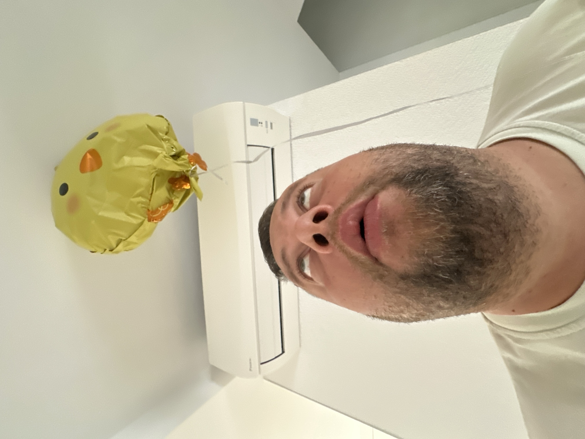 A man, looking upwards with his mouth slightly open, is pictured in a room, possibly in Sant Feliu de Guíxols, Spain. Above him, a yellow balloon shaped like a chick is floating near the ceiling. An air conditioning unit is mounted on the wall behind him. The man is wearing a light-colored t-shirt. The scene appears to be indoors with white walls and ceiling.