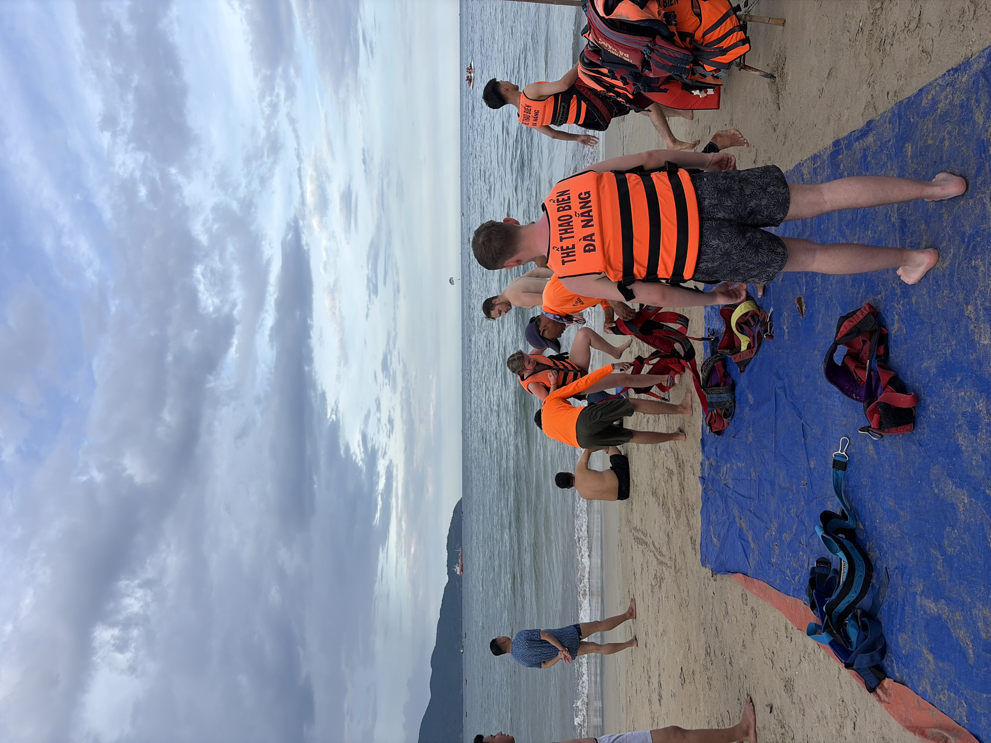 This image depicts a beach scene on a cloudy day, likely in Da Nang, Vietnam, given the text on the life vests. In the foreground, a blue tarp is spread on the sand, with various harnesses and life vests piled on or near it. Several individuals are present, most wearing bright orange life vests, some with text in Vietnamese that translates to "Sports Beach Da Nang."
In the middle ground, people are gathered, some in the process of preparing or assisting with water sports equipment. Two men are standing near the water's edge, and another is seated on the sand. The ocean is visible behind them, with gentle waves breaking on the shore. In the distance, a cargo ship is sailing, and a parasail can be seen in the sky over the water, suggesting activities like parasailing and jet skiing are available. The background features a mountainous coastline. The overall atmosphere suggests a recreational area where water sports are about to commence or have recently concluded.