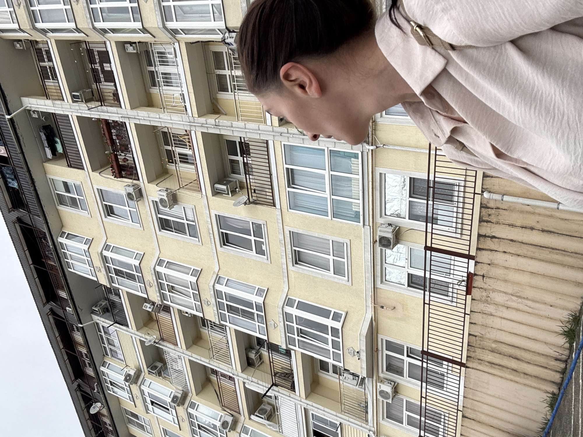 A low-angle, medium shot depicts a person in the upper-right foreground looking down at a large, yellow apartment building in Sochi, Russia. The building features numerous windows, balconies with metal railings, and air conditioning units. The person is wearing a light-colored jacket. The sky is overcast.