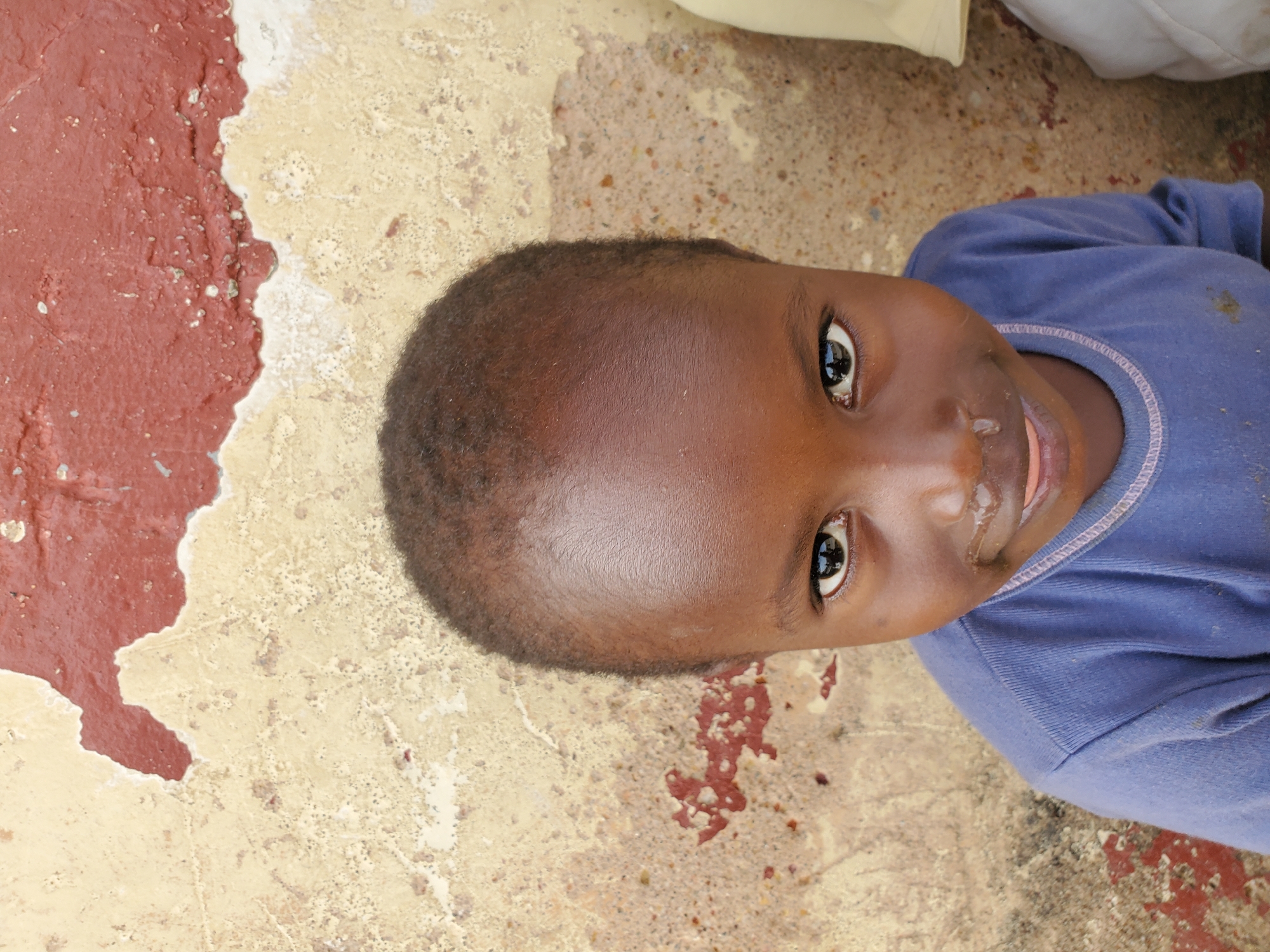 This is a close-up, overhead, outdoor shot of a young child lying on textured ground. The child is dark-skinned and wearing a blue t-shirt. Their head is tilted, and they are looking up towards the camera. The child's mouth is slightly open, and there appears to be some moisture around their lips. The ground beneath them is a rough, mottled surface with patches of red and beige, suggesting it might be a worn pathway or an outdoor play area. There are some small reddish-brown stains scattered on the beige parts of the ground. The lighting appears to be natural daylight. The image composition is dominated by the child's face and the textured ground, with no other significant objects or people in the frame. The overall impression is one of informal observation, possibly capturing a candid moment. The location context is Jos, Nigeria. There is no visible text in the image.
