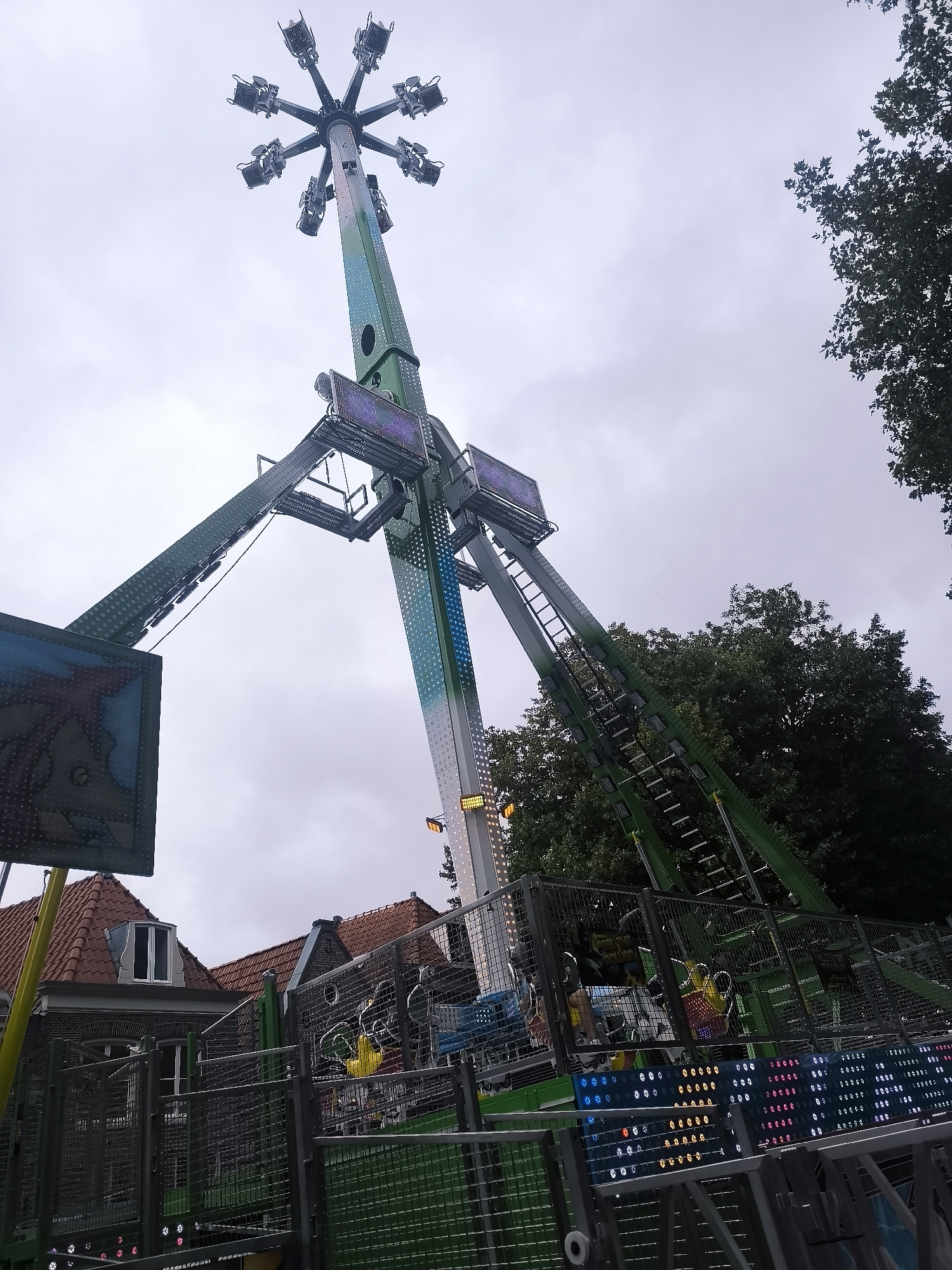 The image shows a large amusement park ride, likely a pendulum ride, situated outdoors. The ride structure is predominantly green and white, with multiple arms extending from a central tower, each holding passenger cars. The ride appears to be stationary. In the foreground and mid-ground, there are sections of metal fencing and what appear to be ticket booths or waiting areas, also with green and grey metallic elements. Some of the fencing has decorative lighting. Behind the ride, red-tiled roofs of buildings are visible, suggesting an urban or suburban setting. Trees with dark green foliage are present in the background, bordering the amusement ride area. The sky is overcast and grey. The scene is located in Hoorn, Netherlands, specifically at the annual fair held at the Veermolen.