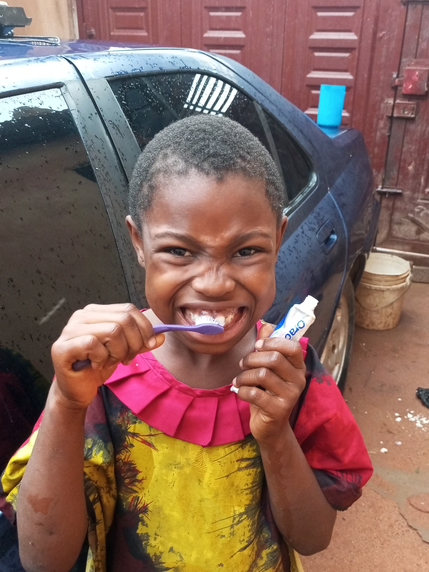 A young Black child with a short haircut grins widely while brushing their teeth with a purple toothbrush and a tube of "Oral-B" toothpaste. The child is wearing a colorful shirt with a yellow and black abstract pattern and a pink ruffled collar. They are standing in front of a dark blue car that is wet with raindrops. In the background, there is a red metal door and a stack of two beige buckets. The ground appears to be wet and made of reddish-brown earth. The scene is set outdoors in the city of Tempure, Nigeria.