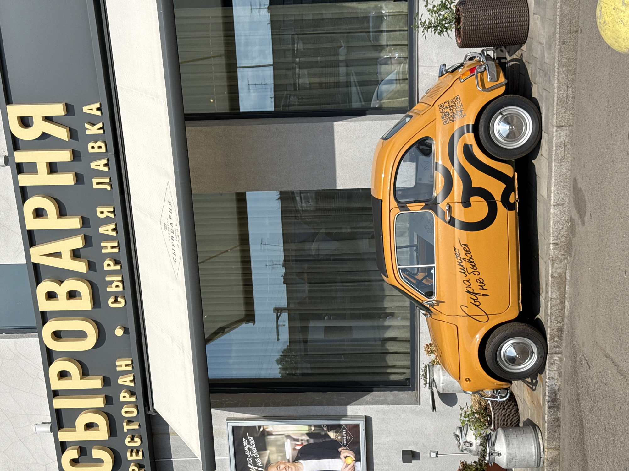 An orange Fiat 500 retro car is parked on a sidewalk in front of a building in Krasnoyarsk, Russia. The building has a large sign with golden Cyrillic lettering that reads "СЫРОВАРНЯ" (Cheese Dairy) above smaller text that reads "РЕСТОРАН" (Restaurant) and "СЫРНАЯ ЛАВКА" (Cheese Shop). An awning extends over a large window of the building. The car has a black graphic design on its side and a QR code on the rear fender. The sidewalk is made of grey concrete and cobblestones. To the left of the car are several large metal milk cans and potted plants. To the right is a large brown wicker planter with a green shrub. A poster is visible on the wall to the left of the car. The weather appears to be sunny.