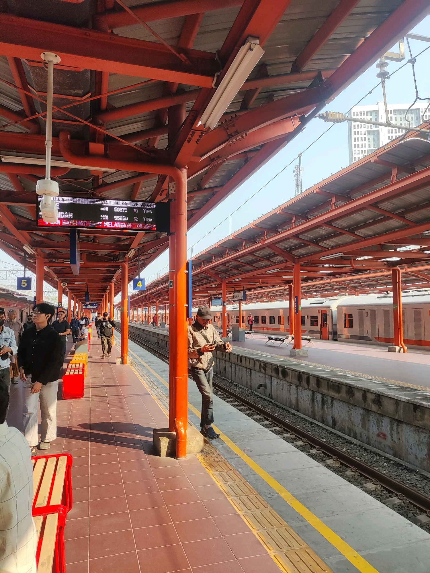 The image shows a view of a train station platform in Bekasi, Indonesia. In the foreground, a tiled platform is visible, with red benches placed along the side. Several people are walking or standing on the platform, some appearing to be waiting for trains.
In the middle ground, a train is parked at the platform. The train is white with orange accents and has several windows. The platform is covered by a red metal roof supported by orange pillars. A digital display board shows train information.
In the background, tall buildings can be seen, suggesting an urban environment. The sky is clear and blue, indicating it is daytime. The overall scene depicts a busy public transportation hub.