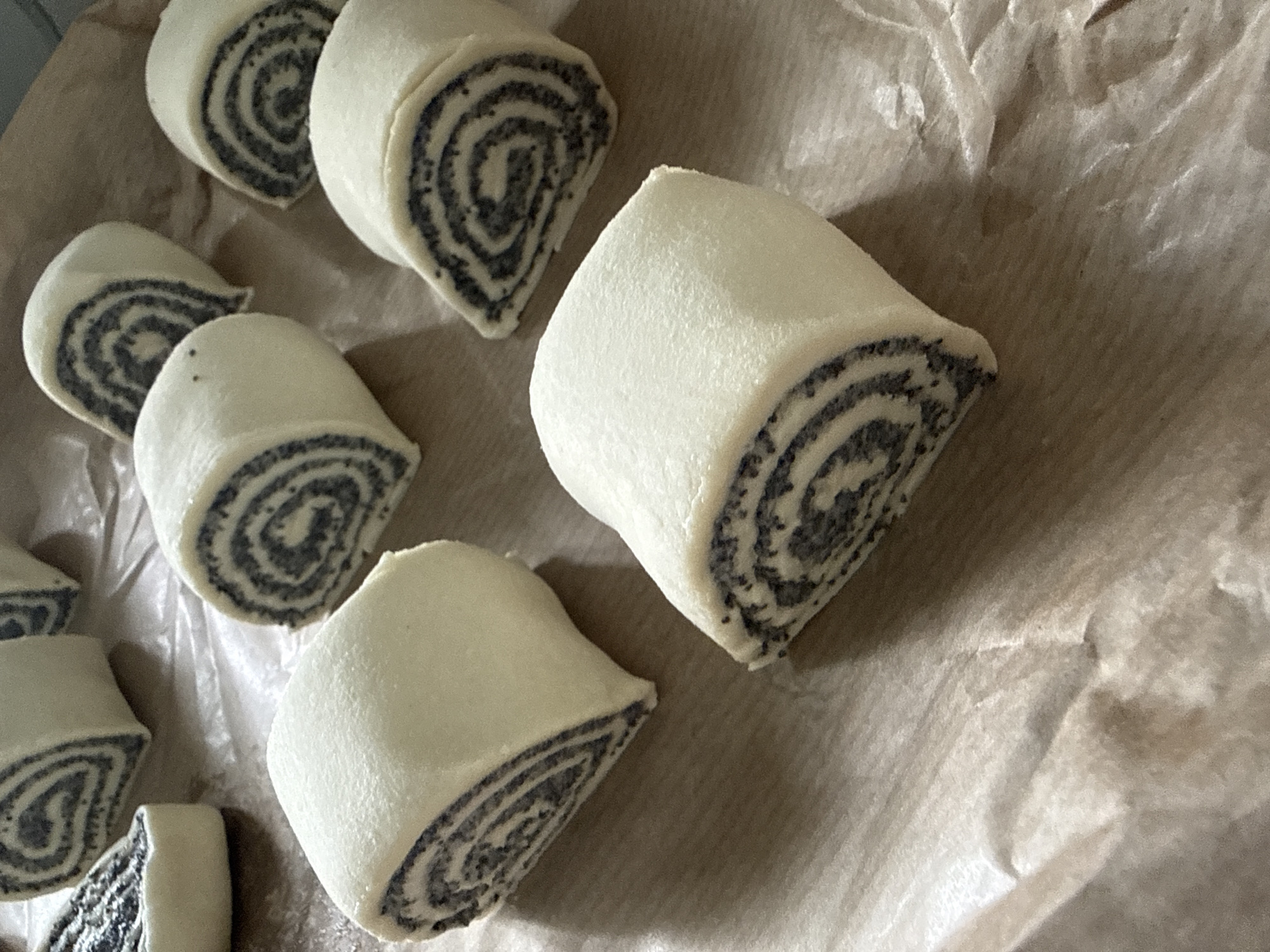 This image displays a close-up, overhead view of several unbaked poppy seed rolls arranged on a piece of parchment paper. The rolls are cut into roughly equal portions, revealing the distinctive spiral pattern of poppy seeds within the pale dough. The parchment paper is laid on a surface that appears to be wood or a similar textured material, with a warm, brown tone. The lighting is soft, creating subtle shadows and highlighting the rounded forms of the rolls. No people are visible in the image, and the scene suggests a domestic or professional kitchen setting, likely in preparation for baking. The image does not contain any information to determine its location in the city of Chaikovsky, Russia.