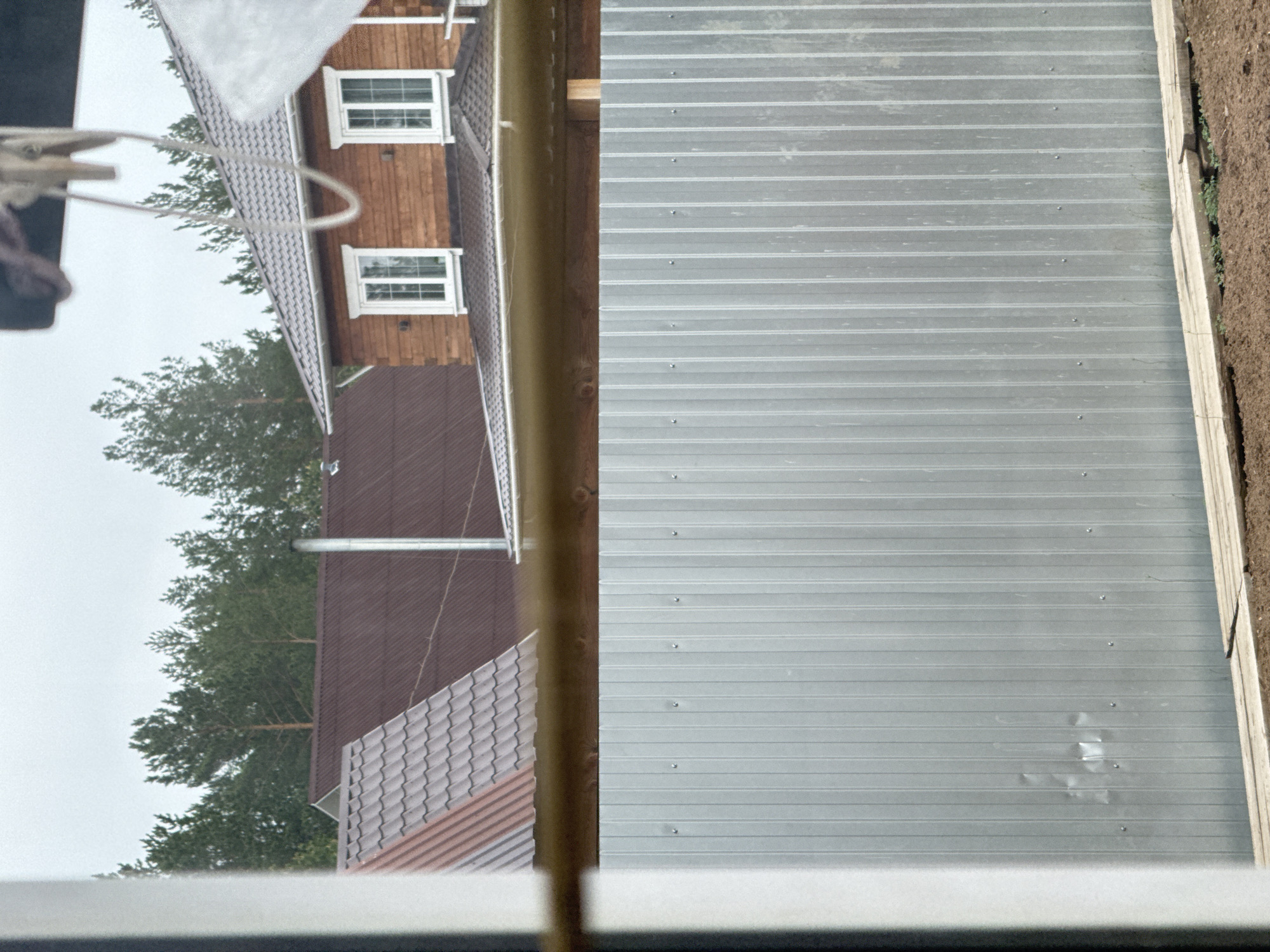 The image shows a view from a window, likely indoors. The window frame is visible at the bottom of the image. Outside the window, there's a view of buildings, trees, and a corrugated metal surface. A wooden building with windows is on the left, next to a roof with tiles. Trees are also seen in the background. A metal sheet, with horizontal lines and rivets is on the right side of the image. The location and specific details about the buildings and environment are not discernible from the image. A clothespin is hanging on the left side of the image.
