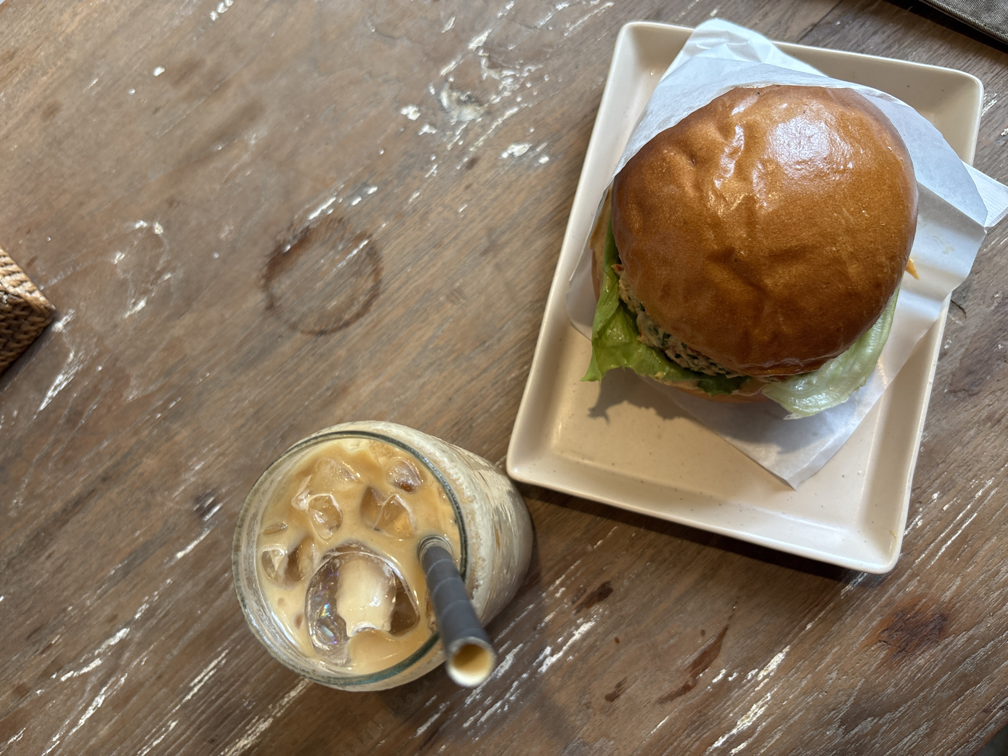 A high-angle, medium shot captures a meal and drink on a worn wooden table. A burger, nestled in white paper on a small, square, off-white plate, sits at the top right of the frame. To the left of the burger is a glass of iced coffee, complete with a straw. A partially visible woven basket is located in the upper left corner of the image. Given the presence of the food items, table, and basket, the setting likely represents a restaurant or cafe, specifically in Kuta Selatan, Indonesia.