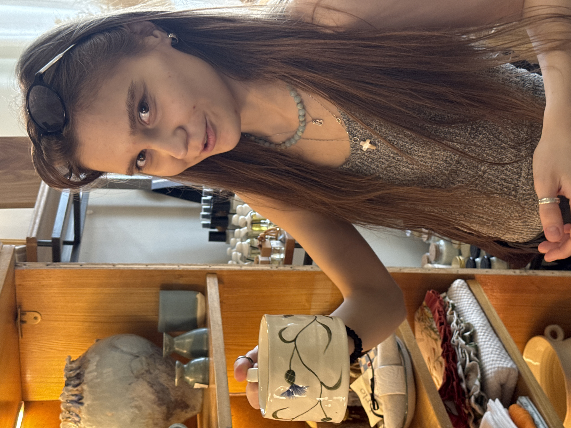 In a dimly lit store, possibly a gift shop or antique store, a young woman with long, light brown hair and sunglasses perched on her head is shown from the chest up. She is wearing a grey, textured tank top and a beaded necklace with a small pendant. In her right hand, she holds a white ceramic jar decorated with blue and green floral motifs. Her left hand is resting near her waist. The background reveals shelves stocked with various items, including ceramic vases, smaller decorative objects, and an assortment of bottled goods, suggesting a retail environment. The lighting appears to be warm and ambient, casting soft shadows. The specific location within Perm, Russia, cannot be determined from the visual information provided.