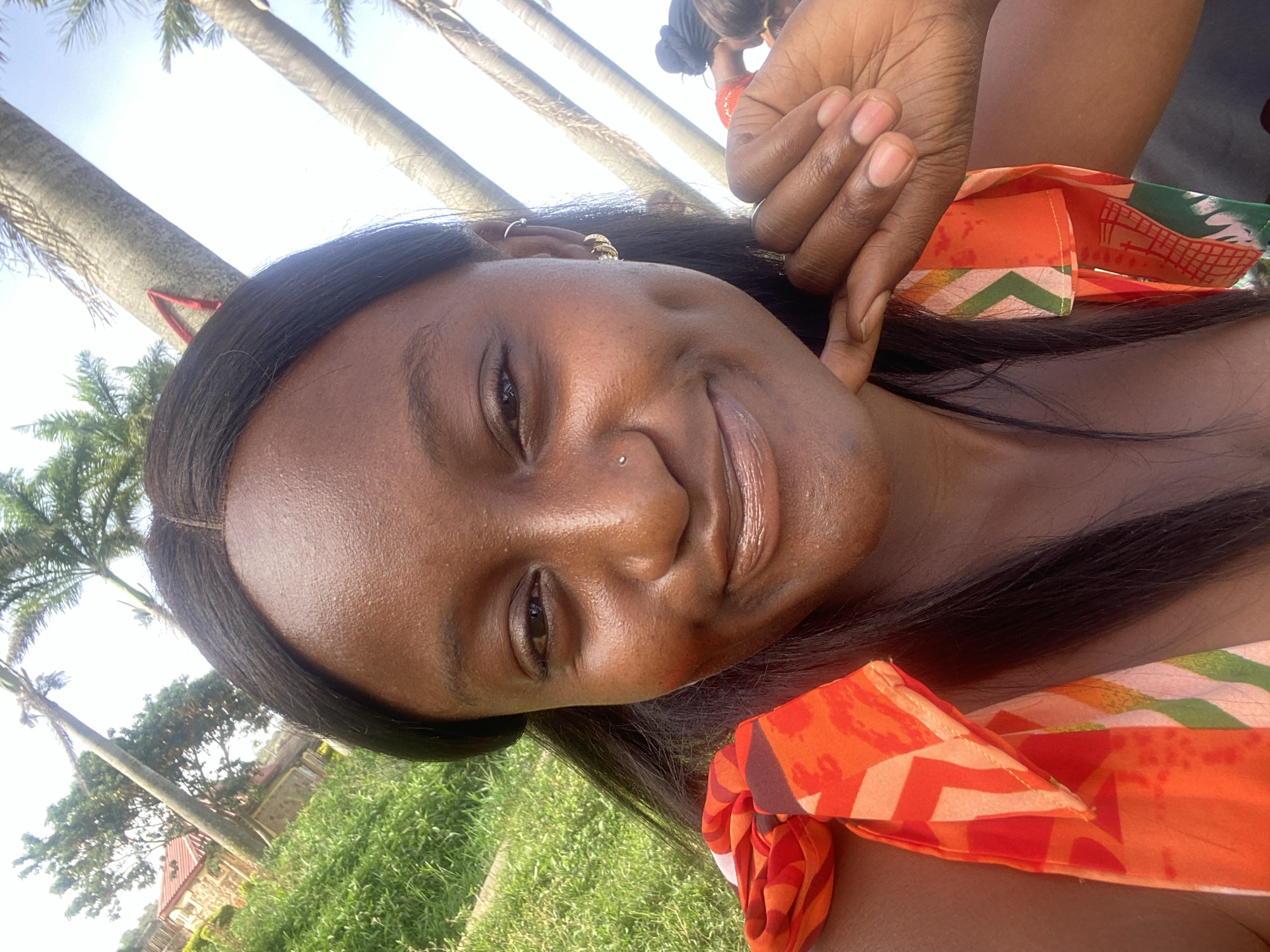 A close-up, slightly tilted portrait captures a young woman with dark skin and long, straight dark hair, smiling warmly at the camera. She has a subtle nose piercing and a small gold hoop earring visible on her left ear, with a dimple appearing on her cheek as she smiles. Her expression conveys happiness and contentment.
Around her neck or shoulder, a vibrant fabric with an orange, green, and white geometric pattern is visible, possibly a headscarf or part of her clothing. In the upper right corner, another person's hands are gently touching near her head, and a glimpse of another individual's head with dark hair styled in a bun can be seen further back.
The setting is outdoors on a bright, sunny day, indicated by the clear, light sky and strong illumination on her face. The background features several tall palm trees and abundant green grass and foliage, suggesting a tropical or subtropical environment. A distant building with a reddish roof is partially visible amidst the greenery. The overall atmosphere is cheerful and relaxed, depicting a pleasant outdoor moment in what is identified as Jos, Nigeria. No visible text is present.