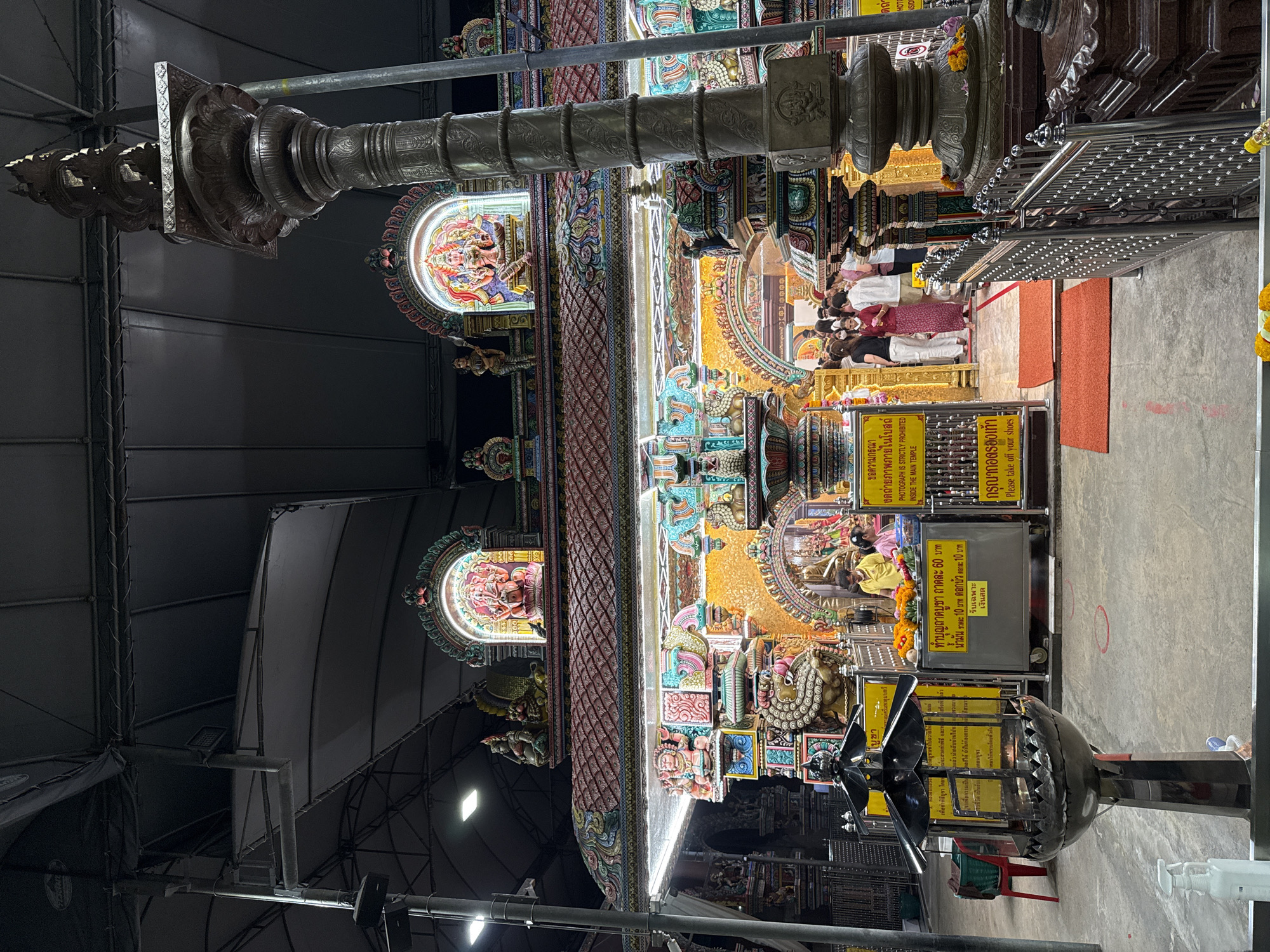 This indoor shot shows the brightly decorated interior of what appears to be a Hindu temple, possibly the Sri Maha Mariamman Temple in Bangkok, Thailand. The temple's ornate architecture features statues and carvings illuminated by artificial light. The scene is viewed from a low angle, looking upwards. People are gathered near the front of the temple. Yellow signs with Thai and English text give instructions to visitors about taking off shoes and no photography inside the main temple. There are payment instructions related to the temple service. Metal structures are visible above, giving some indication of the building's supports. A foreground pillar or structural element partially obscures the view, adding depth to the composition.