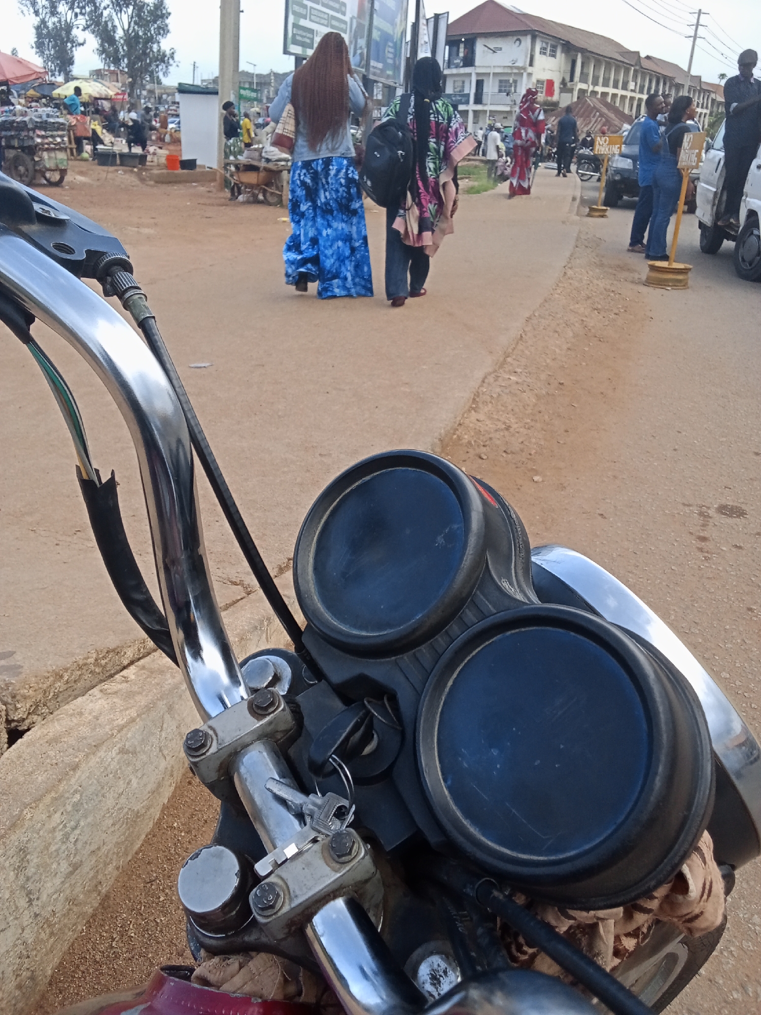 This media file depicts a street scene in Gura Top, Nigeria. The perspective is from someone on a motorcycle, looking forward. In the foreground, the handlebars and dashboard of the motorcycle are visible, featuring two prominent circular gauges and a key in the ignition.
The background shows a bustling street with several people and market stalls. On the left, a woman with long, dark braided hair walks away from the camera, wearing a blue and white patterned skirt and a light-colored top. She is carrying a handbag. Further back, there are market activities with vendors and customers.
To the right of the woman in blue, another woman walks away, wearing a colorful, patterned cloak over jeans and carrying a backpack. Further down the street, several people are walking and interacting. One man is standing on the back of a white vehicle, and another white vehicle is parked with a "NO PARKING" sign in front of it. Buildings line the street in the background.
The scene appears to be during the daytime, with an overcast sky. The ground is dusty and uneven, typical of a street in a developing area. The overall atmosphere suggests everyday life and commerce in a Nigerian town.