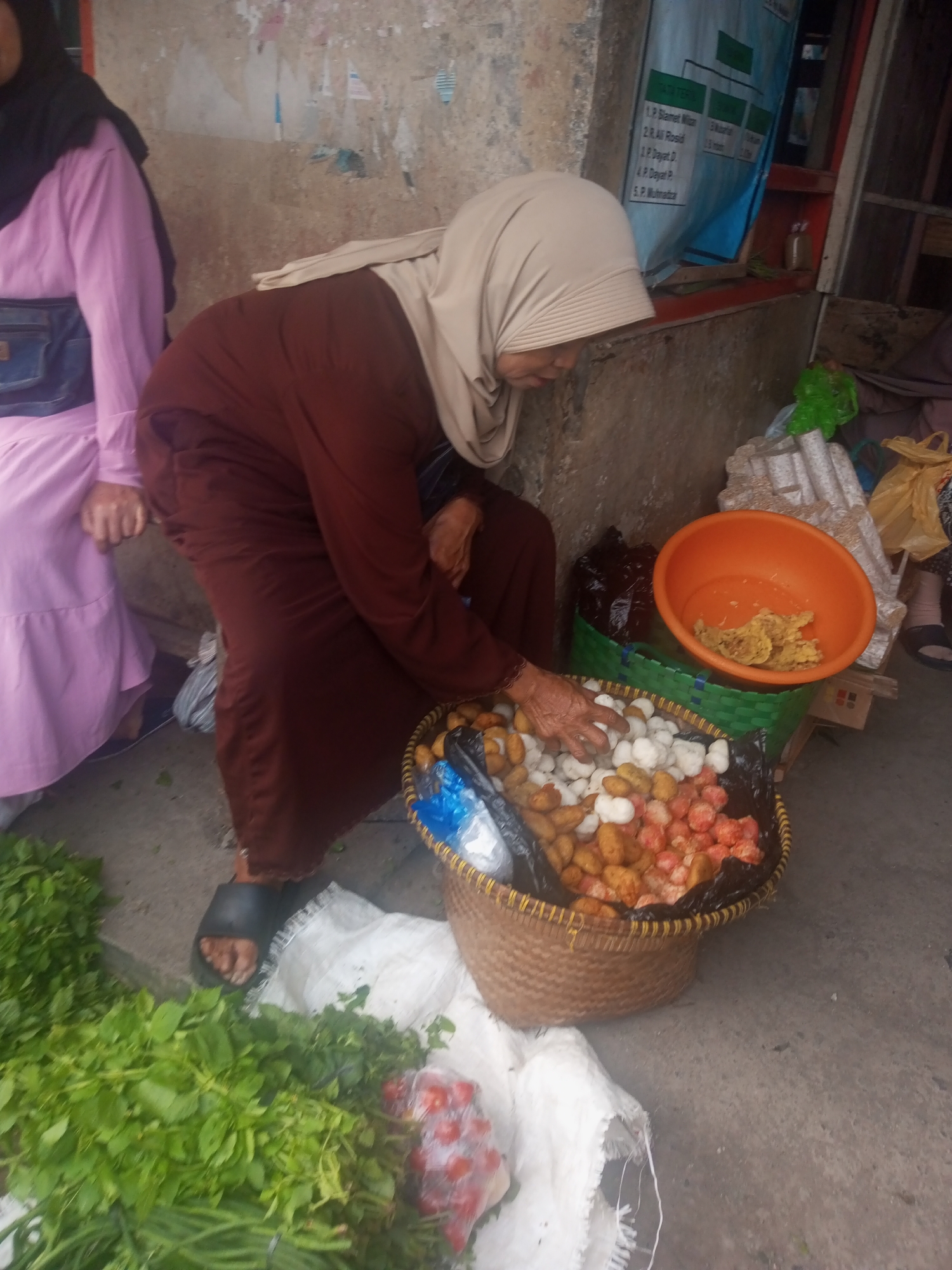 In the city of Kalibeber, Indonesia, an elderly woman wearing a light-colored hijab and a dark brown abaya is crouched down, sorting through a basket of small, round food items. The basket, made of woven straw, is lined with black plastic. The food items appear to be a mix of light-colored and orange-hued morsels. To the left of the basket, a large pile of fresh green leafy vegetables is visible, partially covering a white sack. Another person, wearing a pink hijab and a light purple dress with a dark blue fanny pack, is seated to the far left, their hands resting on their lap. In the background, a rough, textured wall with faded posters is visible, as well as a green woven basket holding an orange bowl containing a yellow, fried food item. A dark brown bag is also visible behind the food basket. The ground appears to be made of concrete.