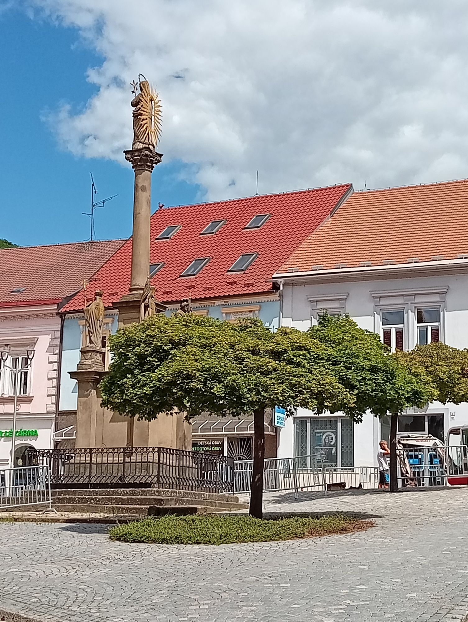 This image shows a view of the town square in Tišnov, Czechia, featuring a prominent Marian column. The column is topped with a gilded statue of the Virgin Mary, radiating light. At the base of the column, two stone statues of saints holding staffs are visible. The column is surrounded by a decorative iron fence and steps leading up to it. In the foreground, a paved cobblestone square is visible, with several well-maintained, round trees planted in front of buildings. The buildings have red tile roofs and white or pink facades with traditional windows. In the background, a blue sky with white clouds is visible. One of the buildings has signs indicating shops, including one for "Dětská Obuv" (Children's Shoes). A woman in a white shirt and blue shorts is standing near a kiosk in the background, and what appears to be a parked truck or bus is also visible. The overall scene suggests a sunny day in a well-maintained European town square.