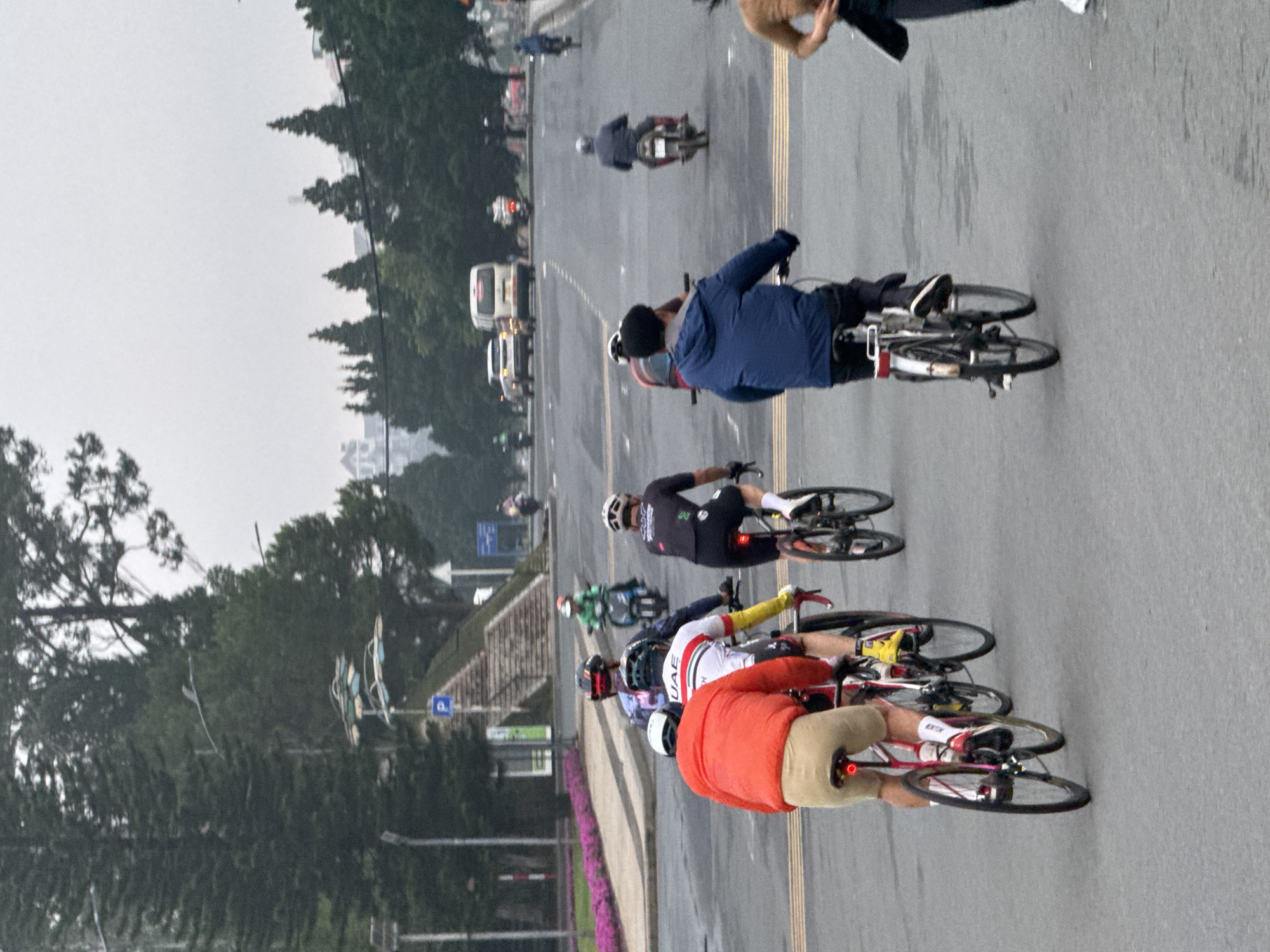 The image depicts a street scene in Phường Xuân Hương, Đà Lạt, Vietnam, likely during daylight hours given the ambient light. Several individuals are engaged in cycling activities on a paved road with lane markings. In the foreground, a cyclist wearing a red jacket and tan shorts is on a bicycle. To their right, another cyclist in black cycling attire is visible. Further back, a group of cyclists are moving away from the camera, one wearing a white jersey with "UAE" printed on it. Several motorcycles and scooters are also present on the road, some with riders. In the background, trees line the street, and buildings can be seen in the distance. The sky is overcast, suggesting potentially rainy or hazy weather conditions. A pedestrian bridge or staircase structure is also visible on the left side of the image, adjacent to the road.