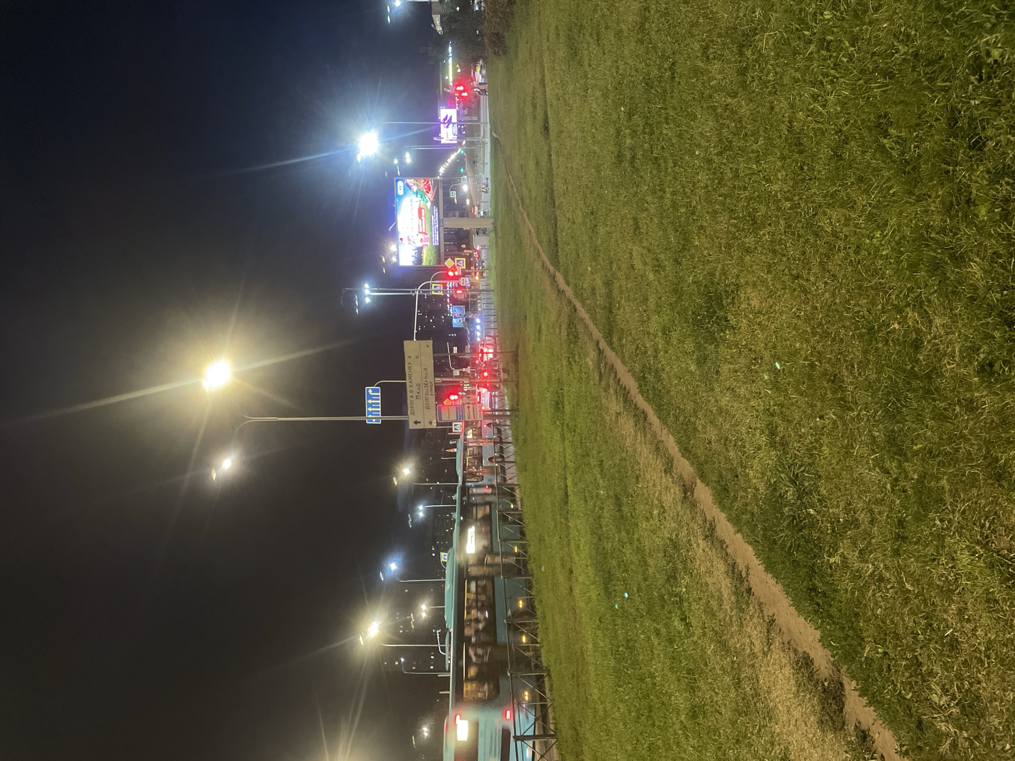 The image depicts a nighttime scene on a city street in Saint Petersburg, Russia, likely an intersection with significant traffic. The foreground is dominated by a grassy median with a worn dirt path. To the left, a turquoise bus with illuminated windows is moving, suggesting active public transportation. A metal fence separates the road from the grassy area.
The background features a busy road with multiple lanes of traffic. Numerous streetlights illuminate the scene, creating glare and lens flares. Traffic lights are visible, with several showing red, indicating a standstill or slowdown. Billboards with bright advertisements are prominently displayed above the road, including one with a picture of a woman and children. The sky is dark, characteristic of nighttime. The overall impression is of an urban environment at night, with artificial lighting and the movement of vehicles being the most prominent features. The architecture and the script on the road signs indicate a Russian context, specifically Saint Petersburg.
