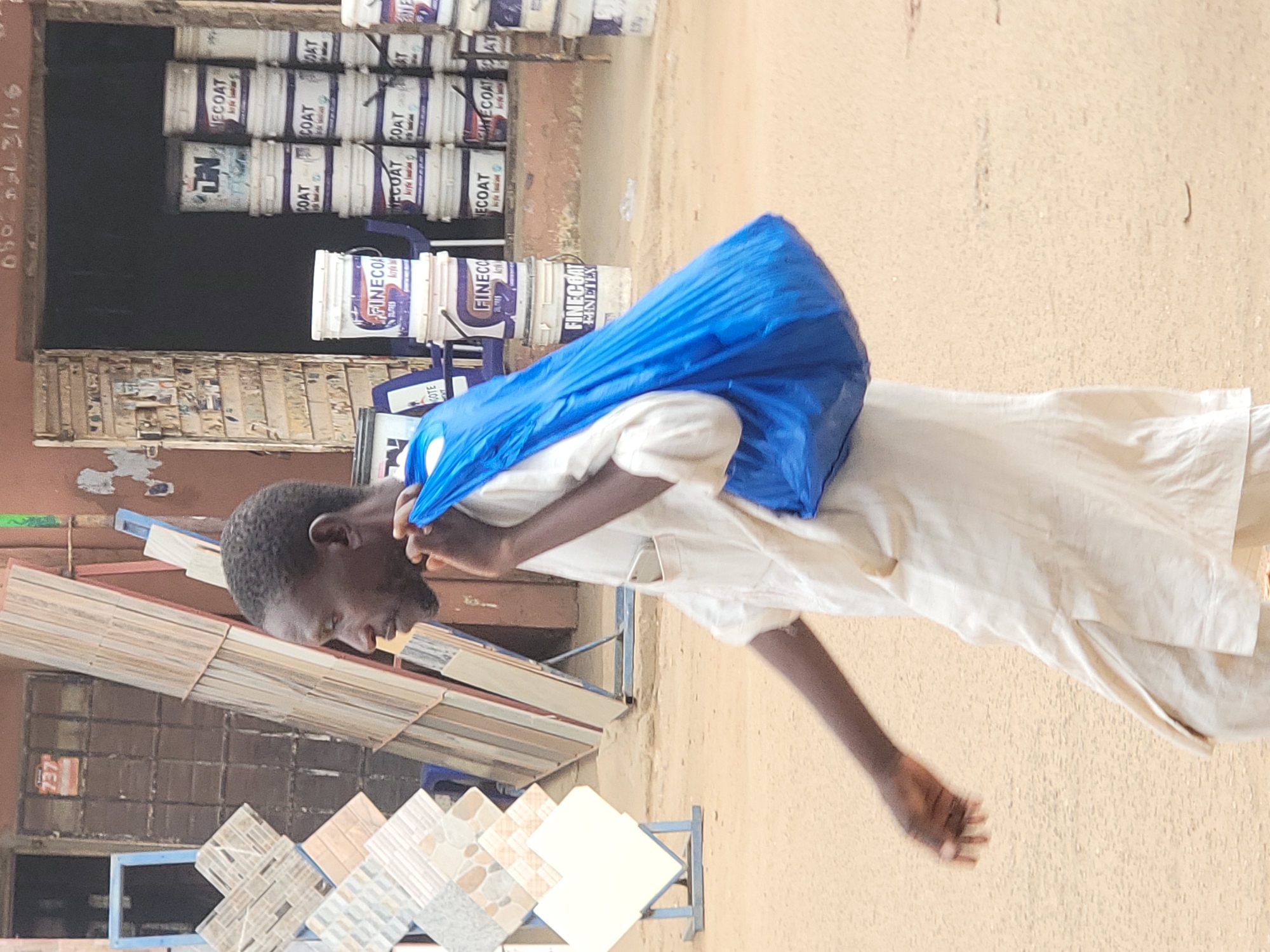 This media file captures an outdoor scene, likely a street or market in Maiburiji, Nigeria, focused on a man walking past a building material shop. The man, a primary subject, is of dark complexion, with short, dark hair and a beard, and is wearing a loose-fitting, light-colored (possibly white or off-white) traditional garment. He carries a vibrant blue plastic bag over his left shoulder, which appears to hold contents.
In the background, a variety of building materials are displayed for sale. Numerous white paint buckets, stacked in rows, are prominent, bearing visible text including brand names such as "FINECOAT," "ECOAT," "COAT," "FINETEX," and "LN." On the left side of the frame, several racks showcase different patterns of ceramic or floor tiles, indicating the nature of the business. The ground appears unpaved and dusty.
Visible text includes the number "737" and a phone number in Arabic numerals, "٩٦١٤٠٦٢٢٢٠१०", on a dark surface to the left, possibly identifying the location or a business contact. The bright, even lighting suggests it is daytime with clear weather. The man appears to be in transit, his focus directed downwards as he walks. The overall setting depicts a typical commercial street scene with local trade activity.