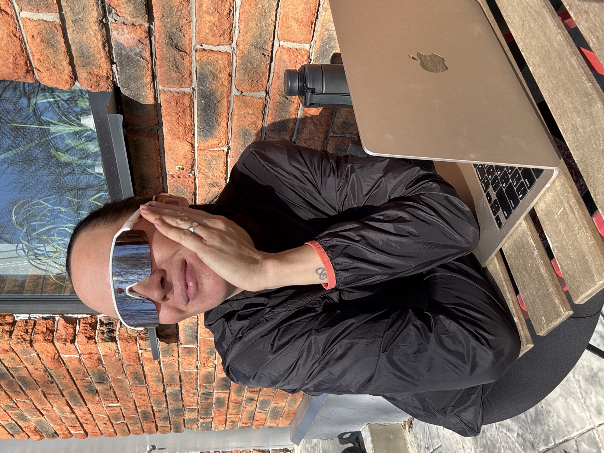 A person wearing oversized white sunglasses and a black jacket is seated at a wooden picnic table. The table is next to a brick wall with a window reflecting trees. A closed Macbook laptop sits on the table, along with a black insulated water bottle. The person's left hand is touching their face and they have a tattoo on their forearm. Based on the brick architecture and presence of trees reflected in the window, the location is potentially an outdoor area in the city of Chaikovsky, Russia.