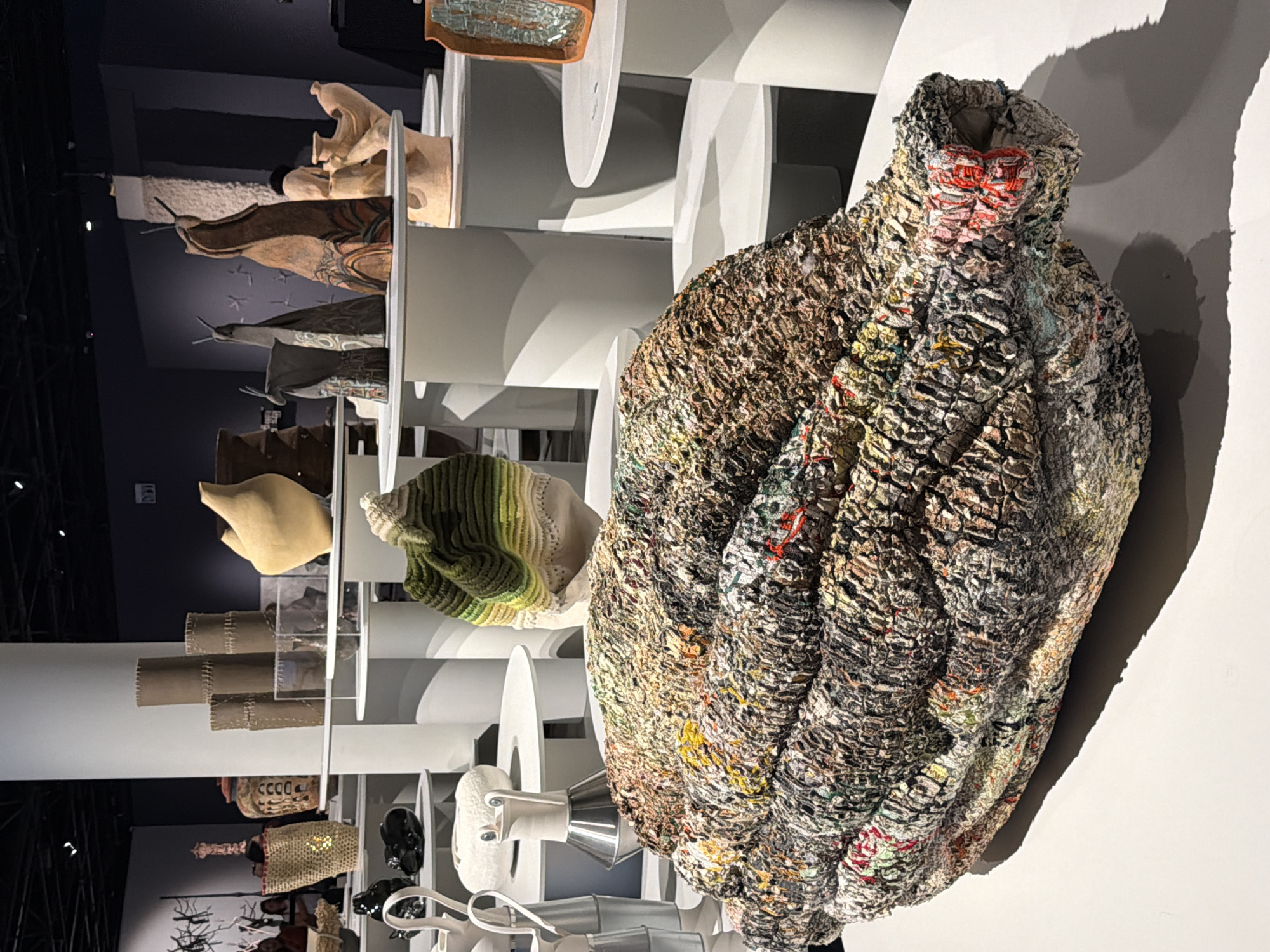 The image shows a piece of textured art, resembling a garment or vase, displayed in an indoor exhibition. The art piece is composed of dense, compacted material in shades of brown, grey, and hints of red, with an uneven surface texture. It is positioned against a white wall and illuminated by a light source from the right, casting a shadow. In the background, a wall-mounted display unit features a series of shelves with other art objects. The image was taken at the Tel Aviv Museum of Art in Tel Aviv, Israel.