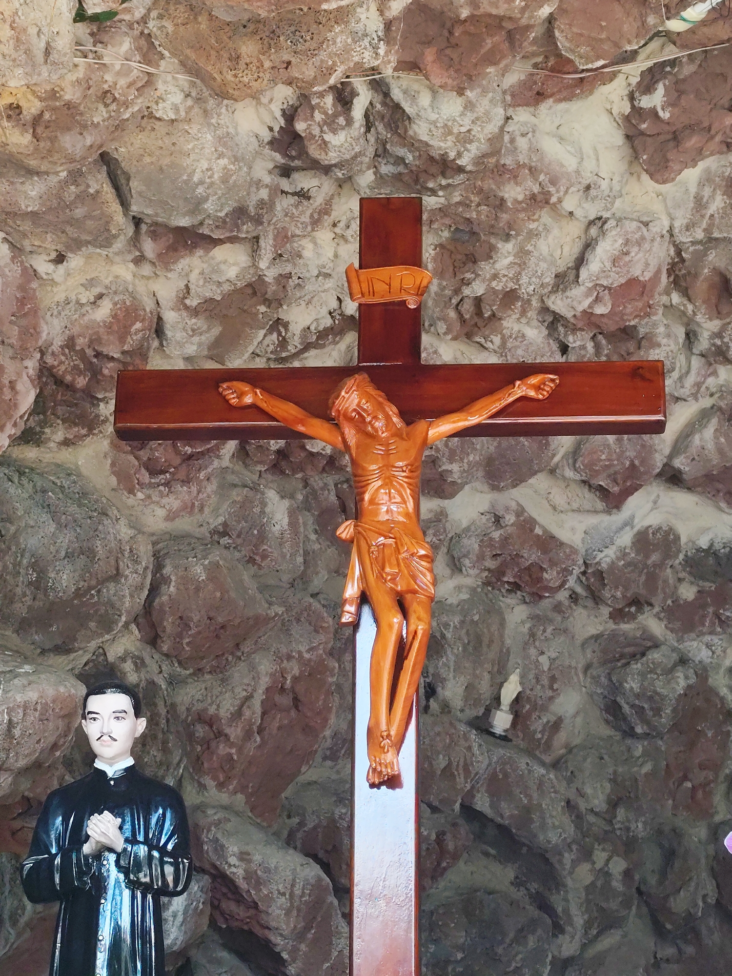 The image shows a statue of a man in a black clerical robe standing to the left of a wooden crucifix with a carved figure of Jesus. The crucifix is positioned in front of a rough, textured stone wall. The man in the statue has dark hair, a thin mustache, and is holding his hands together in front of him. The figure of Jesus on the cross is made of polished wood and has a small plaque labeled "INRI" at the top of the cross. The scene appears to be an indoor or sheltered area, possibly a shrine or chapel, given the religious imagery and the backdrop. This scene is located at the Dinh 2 Palace, also known as the Palace of the Governor-General of Indochina, in Da Lat, Vietnam.