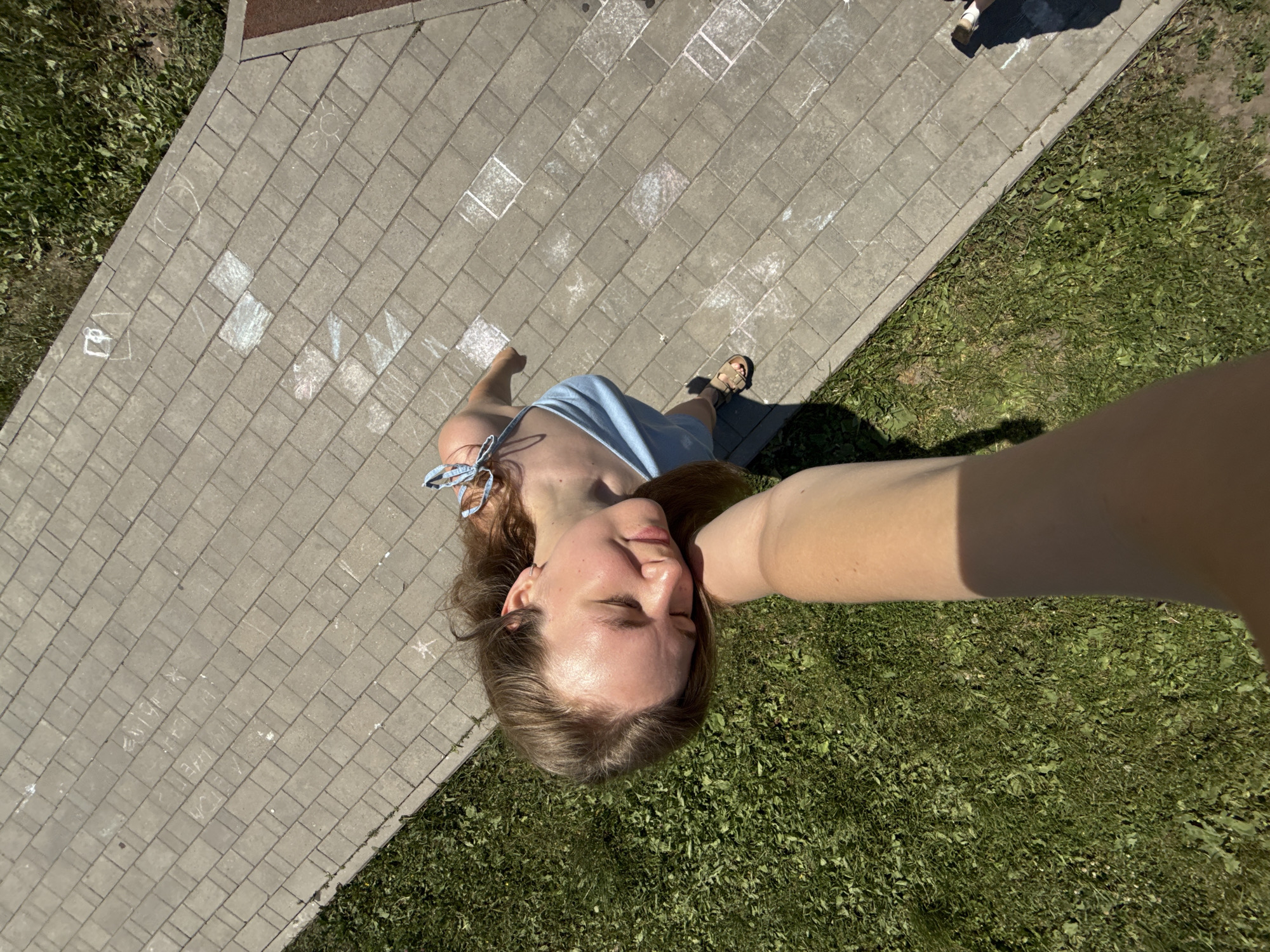 The image depicts a young woman with light brown hair, wearing a light blue sundress and sandals, resting her head on an arm. She is positioned on a paved area adjacent to a grassy section. The paving stones are laid out in a pattern, and some appear to have chalk drawings on them, resembling hopscotch squares or other child-like art. The scene is outdoors, under natural sunlight, suggesting a daytime setting. There are no discernible landmarks to specifically place the location within the city of Vladimir, Russia. The perspective is from above, looking down at the woman.