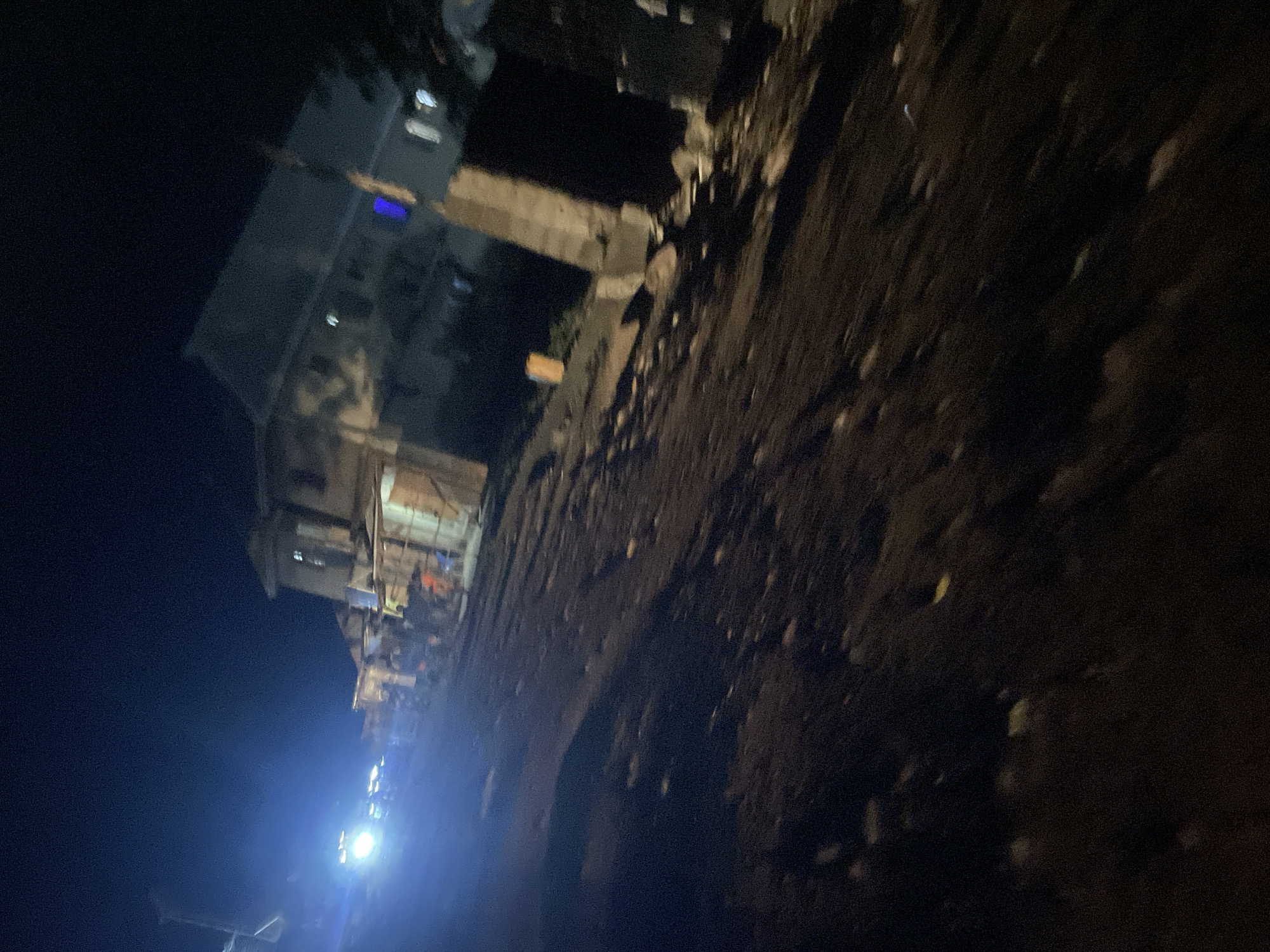 The image captures a nighttime scene on a dirt road in Lokoja, Nigeria. The road is uneven and appears to be unpaved, with scattered debris. On the right side of the road, a multi-story building stands, its windows illuminated. In front of the building, there are several stalls or makeshift structures. To the left, the bright headlights of a vehicle illuminate the road and a group of people are visible, some standing and others possibly engaged in activity. The overall impression is a busy, albeit dimly lit, street scene in a Nigerian city at night.