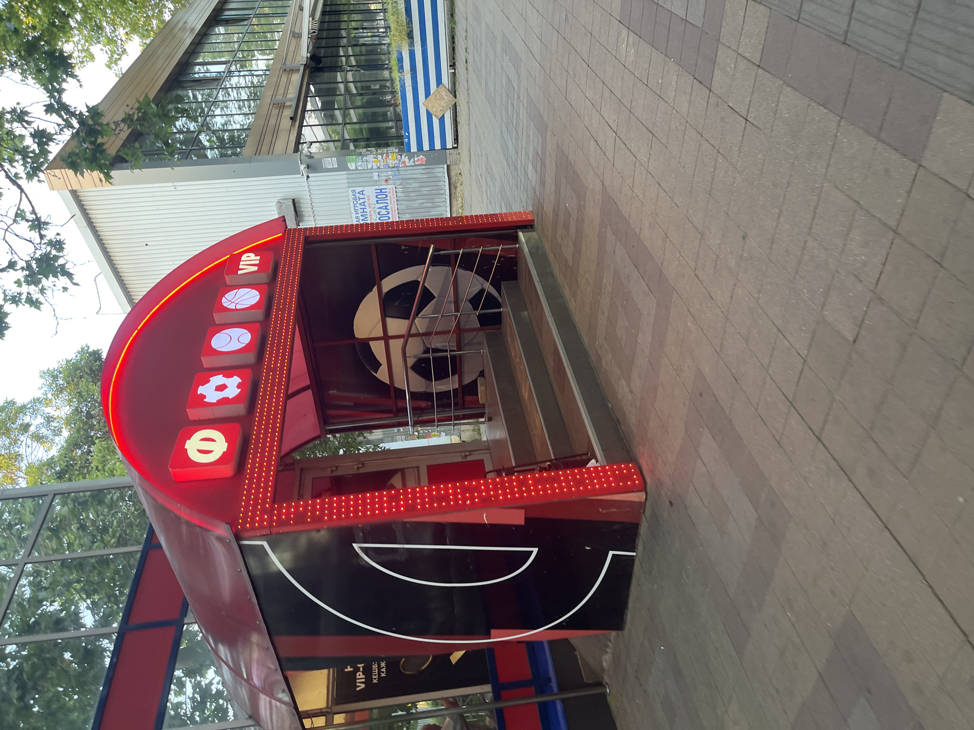 An exterior shot shows a red, arched entranceway with illuminated signage and decorative lights. Above the entrance are three illuminated square icons: a white circle with a symbol, a white gear, and a white outline of a tennis ball. To the right of these icons is a "VIP" sign. The entrance leads to a dark interior with steps and a large soccer ball graphic on the wall. The building to the right of the entrance has a metallic facade and large windows. In the background, trees and a building with a glass facade are visible. The ground in front of the entrance is paved with brick-like tiles. On the left side of the entrance, there is a large black graphic with a white outline, possibly a stylized letter or symbol, and some text that includes "VIP-". The environment appears to be an urban setting.