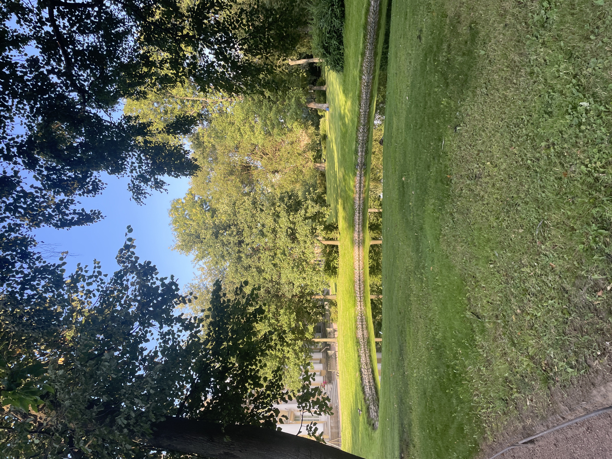 The image shows a serene park scene with a pond in the foreground. The pond has a stone border and reflects the surrounding trees and the clear blue sky. The water is calm, creating a mirror-like surface. The pond is surrounded by lush green grass, which is well-maintained. In the background, there are several mature trees with full foliage, casting dappled shadows on the lawn. To the left of the pond, a light-colored building with white columns and windows is partially visible, suggesting it might be part of an estate or a historical site. A few benches are placed on the lawn between the building and the pond. A person is walking in the distance on the right side of the frame, and a duck is visible near the edge of the pond on the left. The overall atmosphere is tranquil and picturesque. This scene is characteristic of many parks in Saint Petersburg, Russia, such as the Pavlovsk Park or the Catherine Park at Tsarskoye Selo, known for their landscaped gardens and ponds.