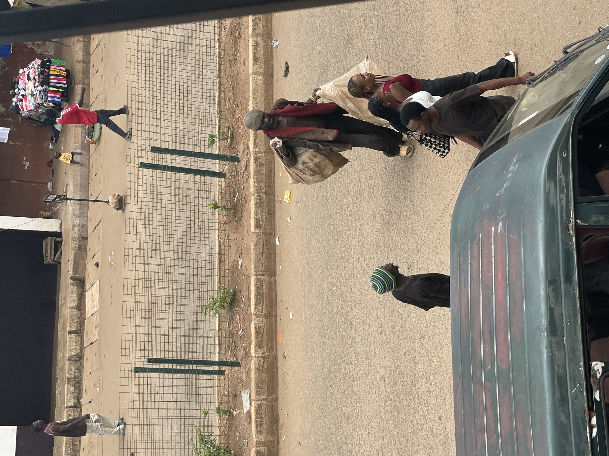 The image captures an overhead view of a bustling street scene. In the immediate foreground, the dark green roof and windshield of a vehicle are visible, with a man in a dark short-sleeved shirt holding its side.
On the main asphalt road, an older man with dark skin, wearing a dark cap, a dark jacket, and a red garment draped across his chest, walks left while carrying two large, light-colored sacks. Walking behind him are two women: one with long dark braids, wearing a white top and carrying a black and white checkered bag; the other with long hair, wearing a red tank top, dark pants, and white sandals. To the left of the vehicle, a man in a black shirt and a green, blue, and yellow striped beanie is partially visible.
Above the main road, a green metal mesh fence separates the street from an elevated dirt embankment with sparse green plants. On a concrete curb beyond this fence, a man in a dark sleeveless top and light pants walks right. Further right, a man in a red short-sleeved shirt and dark pants walks right past a street vendor's stall. This stall consists of a table or cart densely piled with colorful folded garments. A small child in a yellow top is seated on the curb near the vendor's stall. The background features dark building facades and a lighter wall on the far right. The road and dirt surfaces show signs of wear and scattered debris.