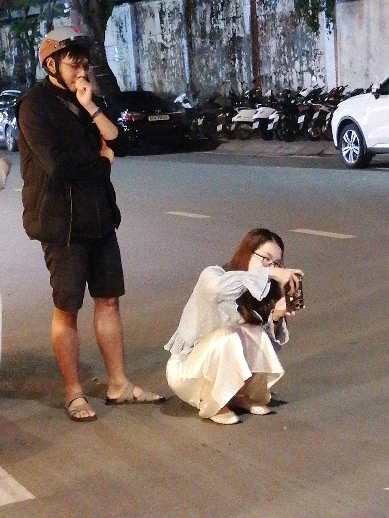 A man wearing a black puffer vest, black shorts, and sandals stands in a street in Da Lat, Vietnam, with his hand to his mouth. Behind him, a woman in a light blue shirt and white skirt crouches and takes a picture with her phone. Motorcycles and a white car are parked in the background. The scene appears to be at night, with artificial lighting illuminating the street.