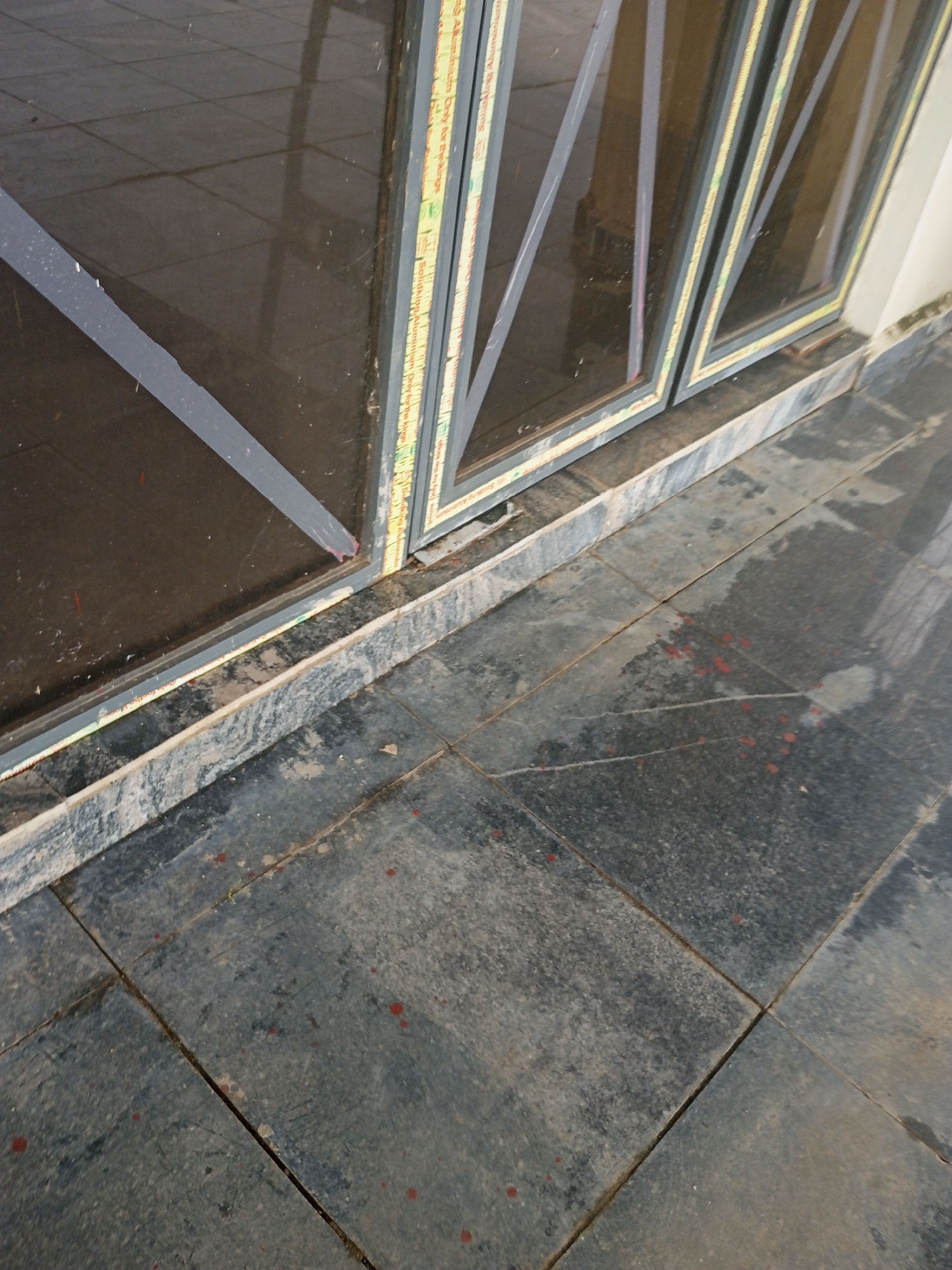 The image shows a close-up view of a window frame and the tiled floor below it. The window is divided into multiple panes by dark gray frames. Some of these frames have diagonal crossbars. A yellow and green sticker or label is visible along the edge of one of the window frames, with some text printed on it. The text is too small and blurry to be clearly read, but some words like "Solid" and "Aluminum" might be present.
The floor is made of dark, rectangular tiles, some of which are cracked or appear worn. There are several small red smudges or stains scattered across the tiles. The floor looks slightly damp or wet in places, and there are reflections on the surface, suggesting it might have rained or been recently cleaned.
The overall scene is outdoors, likely on a ground-level walkway or patio next to a building. The lighting suggests it is daytime, but the specific time of day or weather conditions are not clearly discernible beyond the potential dampness of the floor. There are no people or animals visible in the image. The scene is mundane and shows everyday objects in what appears to be an urban or semi-urban environment in Jalingo, Nigeria. The red smudges on the floor are the most unusual detail.