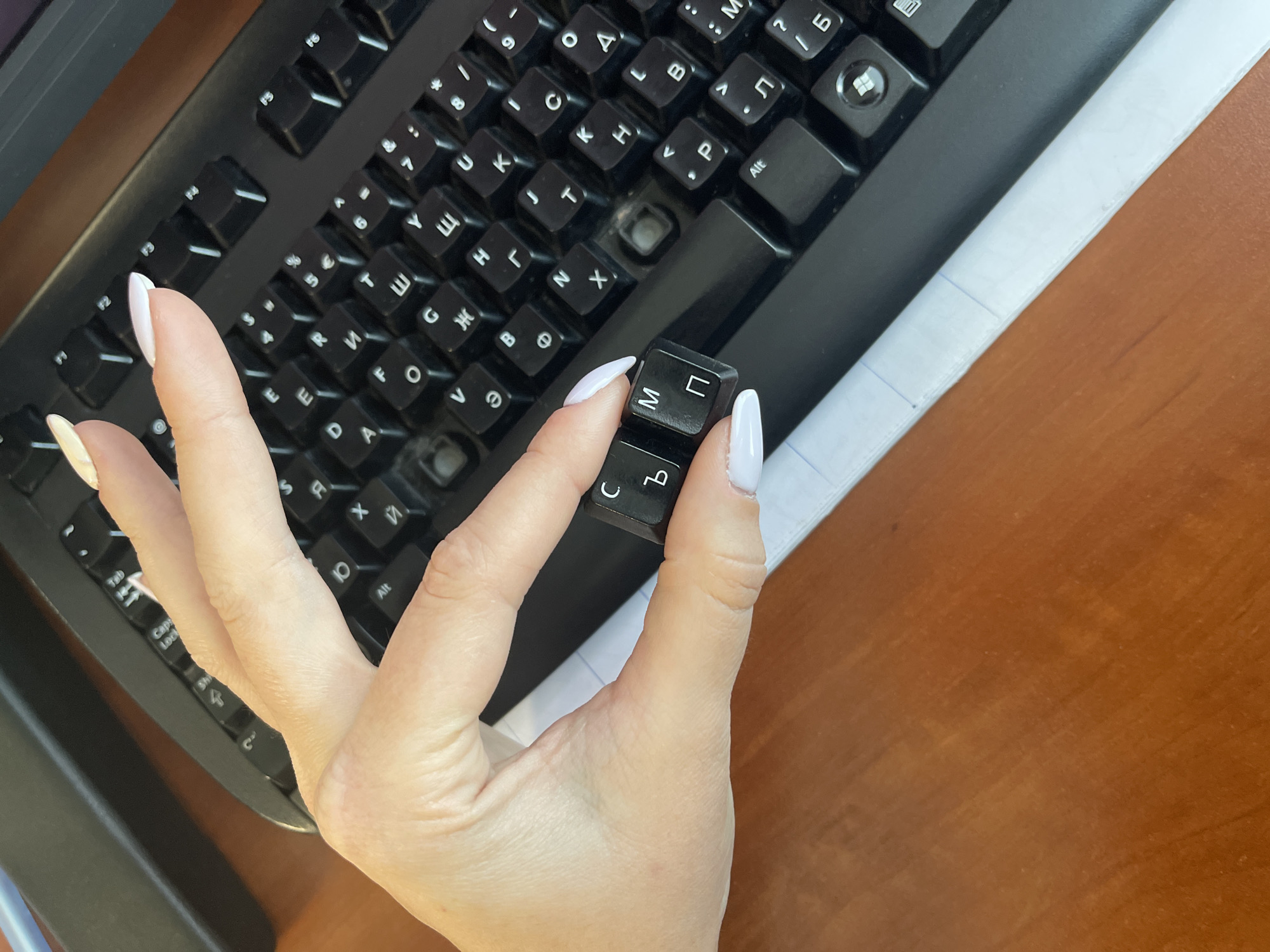 A high-angle shot depicts a black computer keyboard on a brown desk surface, with a sheet of lined paper visible to the right. A hand with long, white manicured nails is holding two detached black keyboard keys displaying Cyrillic characters "M" and "C". The keyboard itself is missing at least one key, leaving a noticeable void. The scene appears to be indoors, possibly in an office or home setting. Since the keyboard keys feature Cyrillic characters, the scene might be located in a region where this script is used, such as Sofia, Bulgaria, though this cannot be definitively determined from the image alone. The overall impression is one of disrepair or maintenance of the keyboard.