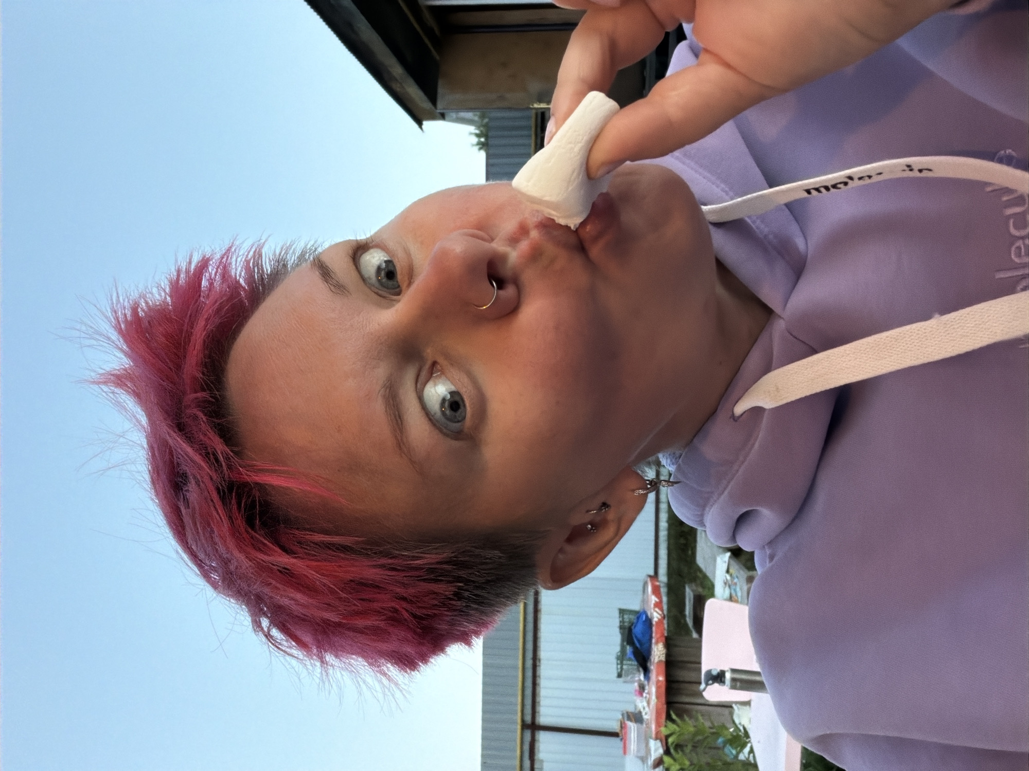 An adult with short, spiky, bright pink hair is captured in a close-up selfie outdoors against a clear blue sky and a blurred background of a building and fence. The individual is wearing a light purple hooded sweatshirt and has a silver hoop piercing in their nose and a silver earring in their left ear. They are holding a white marshmallow to their lips with their right hand, with their mouth puckered as if to eat it. The person's eyes are wide open, and their head is tilted to the left. The scene appears to be in a residential area.