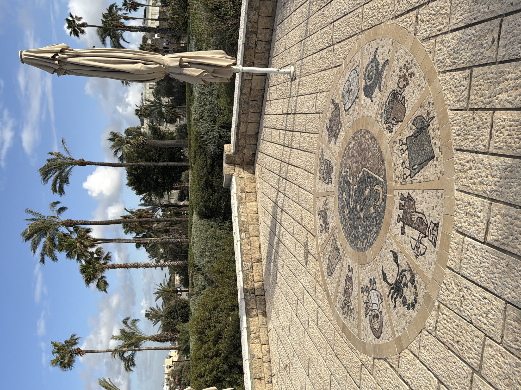 The image shows a mosaic zodiac clock embedded in a stone-tiled walkway in the city of Tel-Aviv, Israel. Palm trees and blue sky with scattered clouds are visible in the background, along with various bushes and greenery. An unopened beige parasol stands closed on the left side of the image. The scene is brightly lit, and the camera angle is slightly tilted.