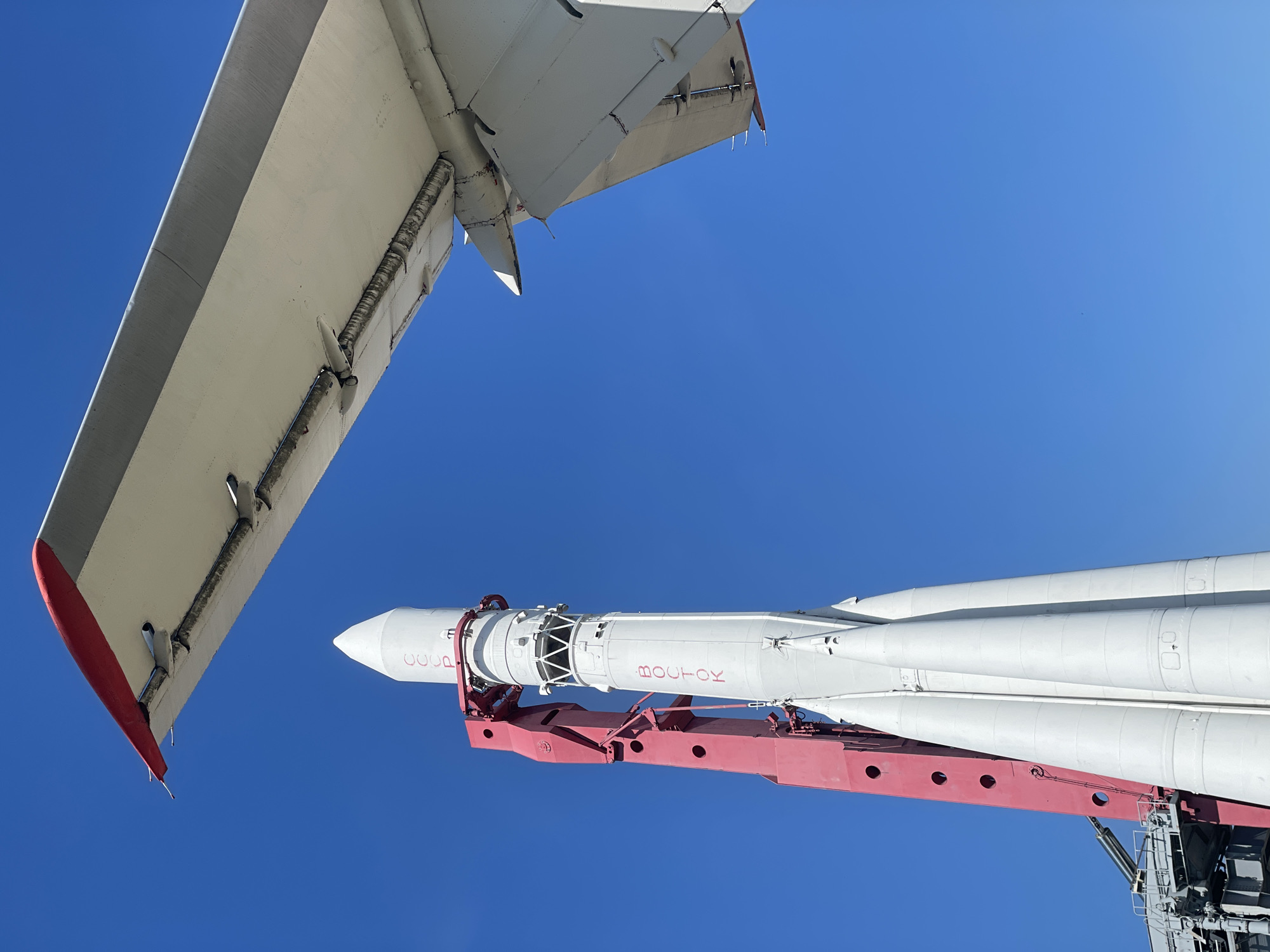 The image shows a low-angle view of a rocket and an aircraft wing against a clear blue sky. The rocket is white with red lettering in Russian, and is mounted on a red support structure. The aircraft wing is white with red accents and appears to be part of a larger aircraft. The scene is likely at a museum or exhibition.