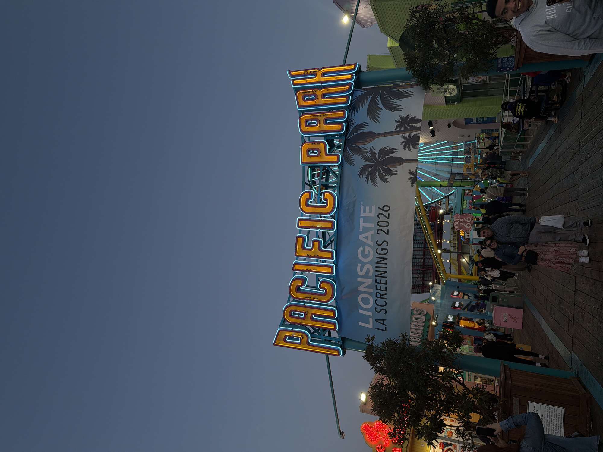 The image shows the iconic "Pacific Park" sign at Santa Monica Pier in Santa Monica, California. The sign is prominently displayed, featuring orange and blue neon lights. A banner beneath the sign reads "LIONSGATE LA SCREENINGS 2026" with palm tree graphics. The Santa Monica Ferris wheel is visible in the background. People are walking on the pier along a wooden boardwalk. Some are tourists. In the foreground, there are food stands and arcade games. The sky is an early evening blue.