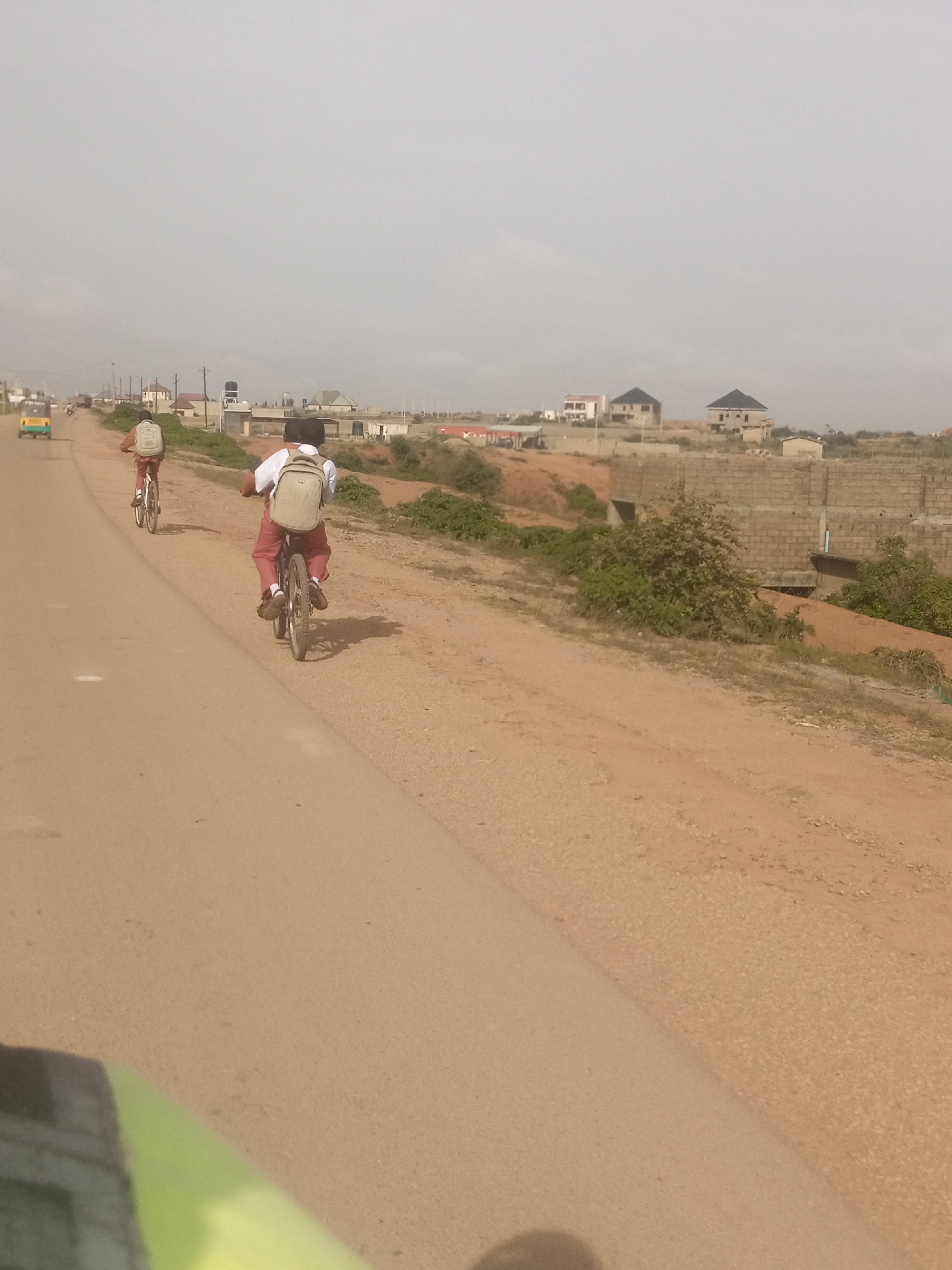 In Rayfield, Nigeria, two individuals are observed cycling on a paved road. The person in the foreground, wearing a white shirt and carrying a light-colored backpack, is riding a bicycle. Behind them, another person in a brown shirt and also with a backpack is cycling. The road is bordered by a sandy, uneven terrain on the right, with vegetation and some brick construction visible in the distance. Buildings, including what appear to be houses under construction, are scattered across the landscape. The sky is overcast. A yellow and green vehicle, possibly a tuk-tuk, is seen further down the road in the background.