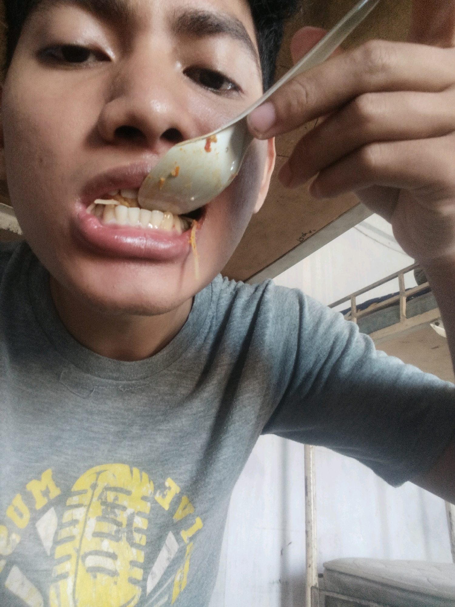 A person is eating with a spoon, holding it to their mouth. They are wearing a gray t-shirt with a yellow microphone graphic. The background appears to be indoors, with a metal frame and some white fabric visible. The lighting is somewhat dim. The image is likely a selfie taken by the person eating. There are no identifiable landmarks or indications of the specific location within Medan, Indonesia.