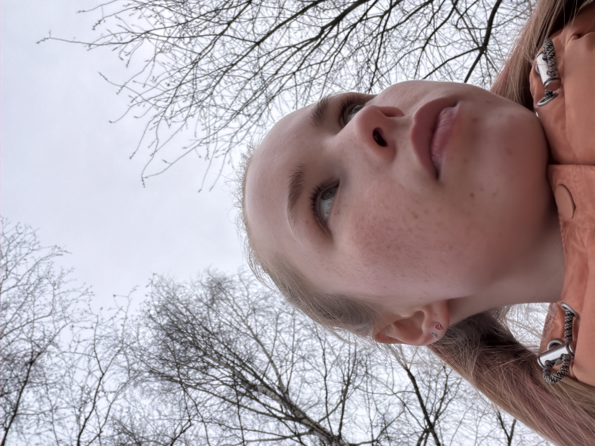 A low-angle, upward-facing photograph depicts a fair-skinned person with light blonde hair, presumably female, with freckles on her face. The person's face is oriented in such a way that the viewer is looking up at them. She is wearing an orange jacket. A tree with bare branches is visible in the background, and the sky is overcast. Location is not discernible from the image.