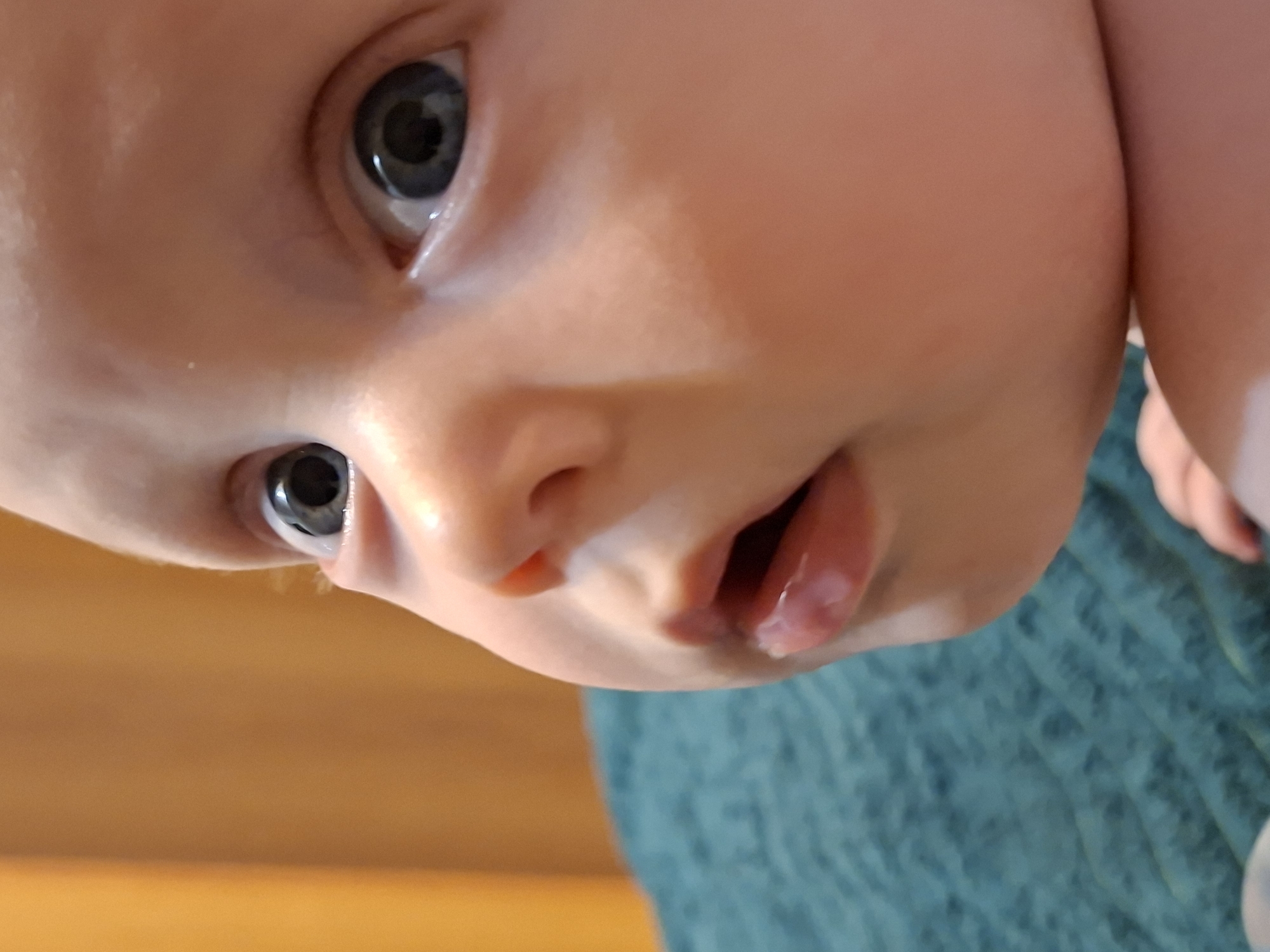 A close-up, upside-down perspective captures the face of an infant with fair skin and large, dark eyes. The baby's mouth is slightly open, with a small amount of drool visible on the lower lip. The baby is lying on a blue, textured fabric. The background suggests the presence of a wooden surface or object, out of focus. The location and city are unknown.