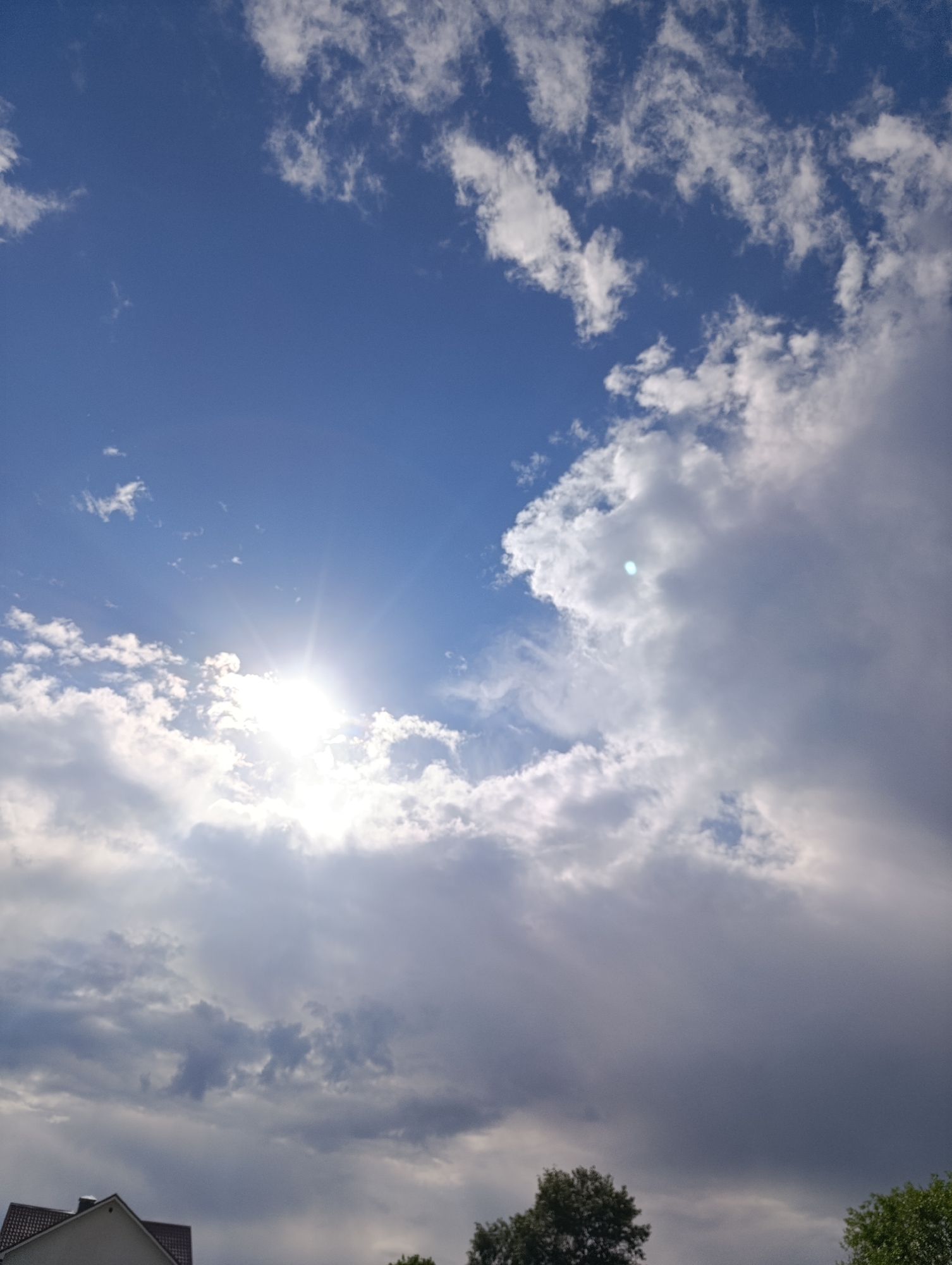 The image depicts a daytime sky in Unknown, Unknown. The sky is predominantly blue, partially obscured by white and grey clouds. The sun is visible, shining brightly through a gap in the clouds, creating a starburst effect. In the lower portion of the image, there is partial visibility of a building with a red-tiled roof and some trees.