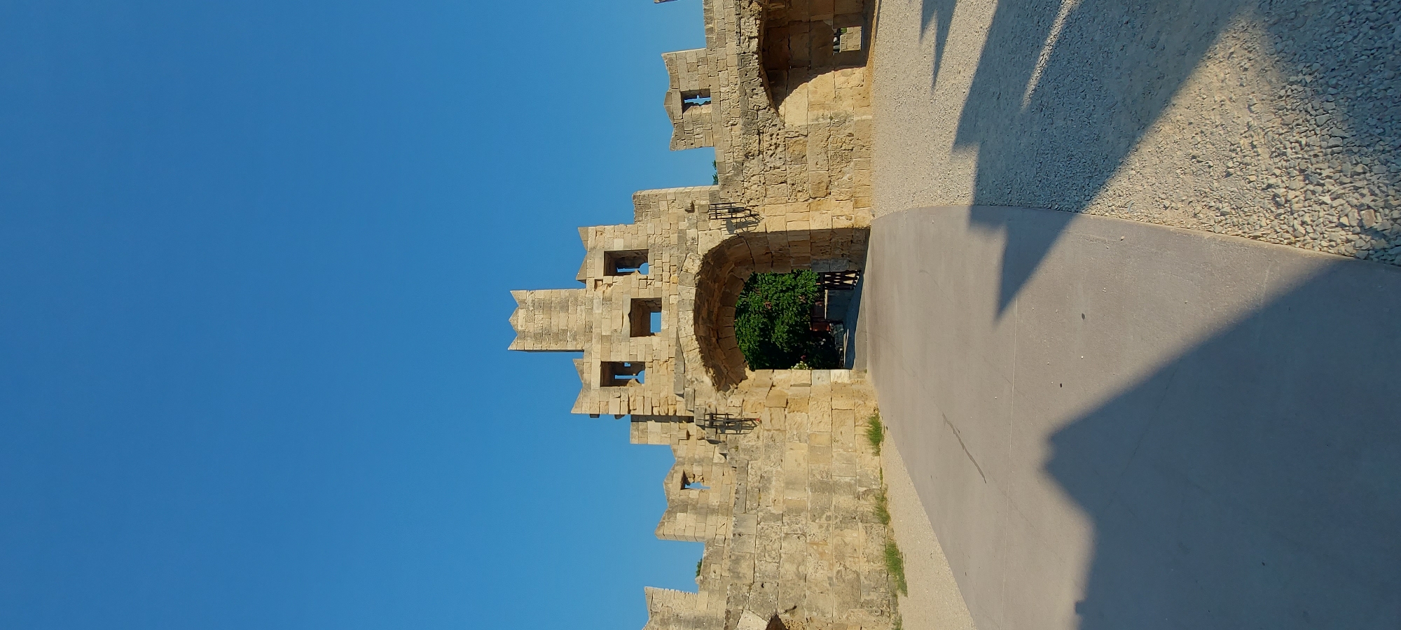 The image shows the Archangel Michael Gate, a medieval stone gatehouse in the city of Rhodes, Greece. The gate is constructed from large, rough-hewn stones and features crenellations at the top, indicative of its defensive purpose. An arched passage passes through the center of the gate, revealing a glimpse of greenery and a wooden fence beyond. The area in front of the gate is paved with concrete and gravel. The sky is a clear, vibrant blue. The sun casts long shadows on the ground, suggesting it is late afternoon or early morning. The overall scene is one of ancient architecture set against a bright, open sky.