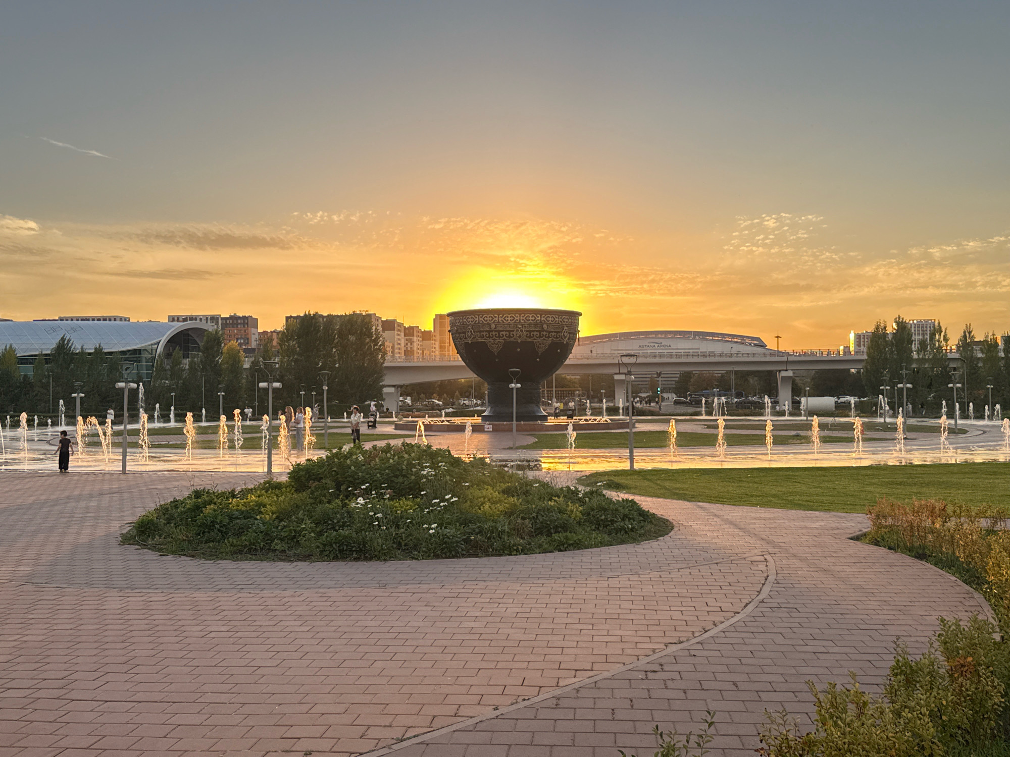 A wide shot captures a park scene in Astana, Kazakhstan, at sunset. The sun, a bright orange orb, is setting behind a large, ornate black bowl-shaped monument in the mid-ground, its light illuminating the sky with warm hues of orange and yellow. The monument, with intricate gold patterns, is surrounded by a circular fountain where water jets create illuminated sprays, catching the golden light. A winding brick pathway dominates the foreground, leading towards the fountain area. Lush green grass and a patch of white-flowered bushes are visible to the right of the pathway and behind the fountain. In the background, modern city buildings, including a stadium labeled "Astana Arena," and an elevated road with cars are silhouetted against the sky. Several people are present, scattered around the fountain area and walking along the pathway. The overall atmosphere is peaceful, bathed in the soft glow of the setting sun.