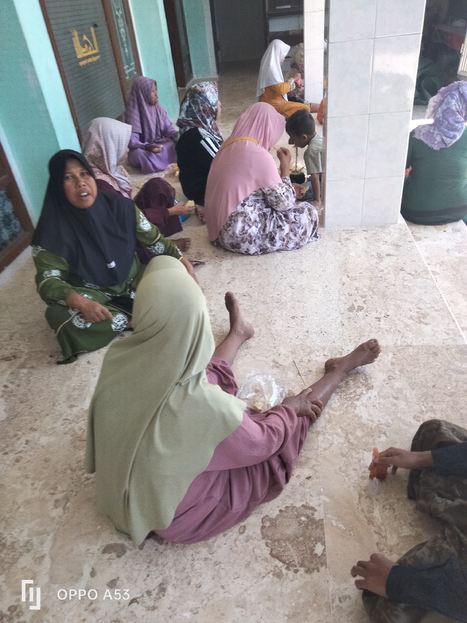 In a communal indoor setting, likely a public space such as a mosque or community center in Badung, Indonesia, several individuals are seated or lying on a tiled floor. The floor is patterned with beige and light brown tiles. The background features a light green wall and pillars with white square tiles. A prominent glass door with a symbol is visible on the left wall.
A woman in a black headscarf and green patterned dress is seated on the floor, looking towards the camera. To her left, another woman in a light pink headscarf sits. Behind them, a woman in a dark purple headscarf and dress is seated, and next to her, another woman wears a patterned headscarf and a black top with white stripes. A young child, wearing a light brown top and dark pants, is being attended to by a woman in a pink headscarf. Further to the right, a person in a green garment with a light purple headscarf is seated facing away from the camera. In the foreground, a person in a light green headscarf and a pink garment is lying down, with their legs extended. Another person's hands are visible in the lower right corner, interacting with a small orange object, possibly food. Plastic packaging is visible near the person lying down.
The atmosphere appears to be one of gathering or waiting, with some individuals engaged in eating or interaction. The lighting is diffused, suggesting an overcast day or an indoor environment without direct sunlight. The presence of people in traditional Islamic attire suggests a cultural context common in Indonesia.