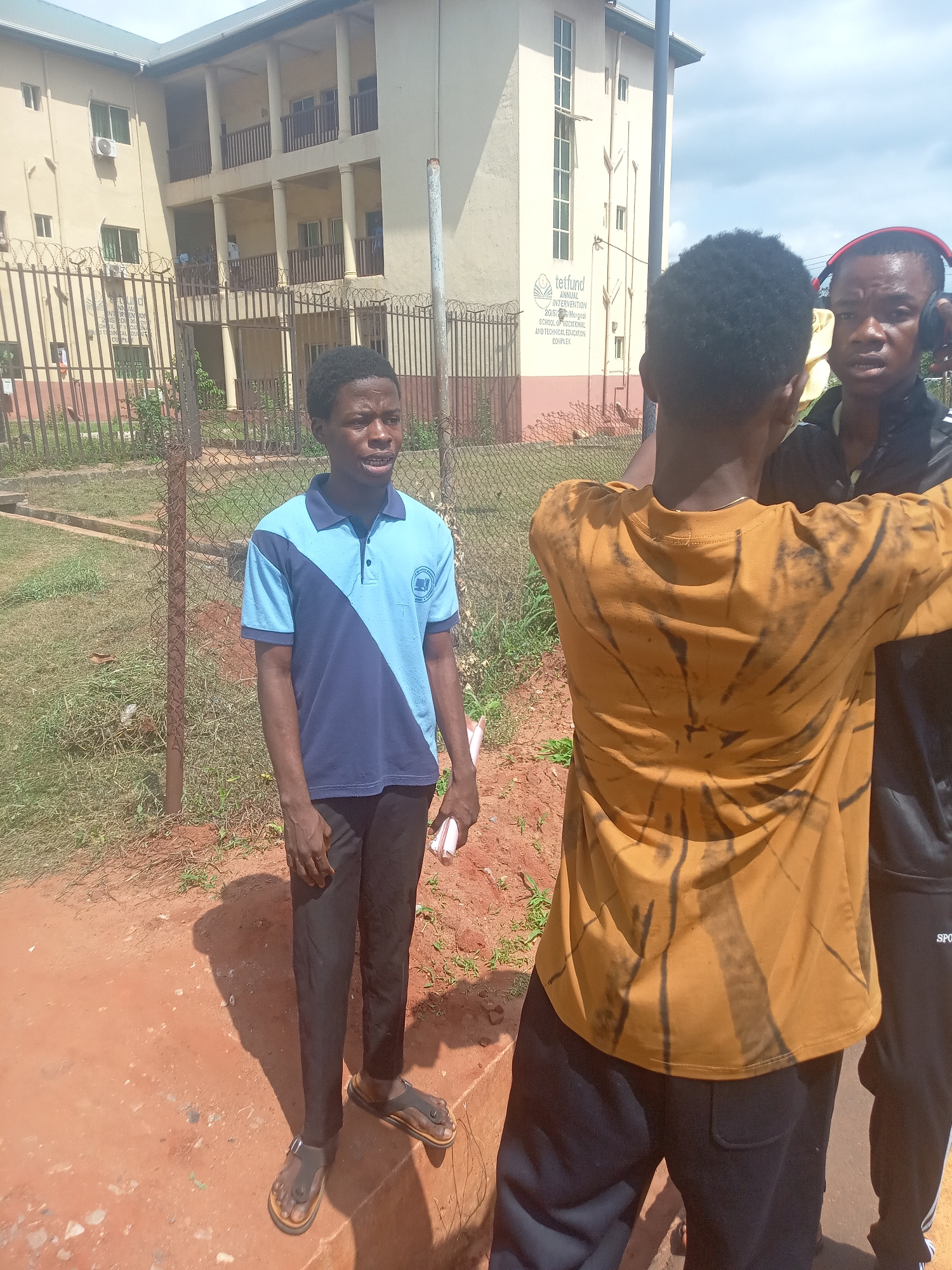 Three young men are captured outdoors in Nsugbe, Nigeria, on a bright, sunny day. The scene features reddish unpaved ground with patches of grass in the foreground. In the background, a multi-story, light-colored building with multiple windows, balconies, and air conditioning units stands behind a metal fence, topped with barbed wire.
On the left, a young man with dark skin and short dark hair wears a two-tone polo shirt (light blue and dark blue), black trousers, and dark sandals. He is seen speaking, looking towards the right, and holding a small white object, possibly paper or a phone.
The central figure has his back to the camera, wearing a mustard yellow or orange t-shirt with dark, abstract tie-dye patterns and dark trousers. He is oriented towards the man on the left.
On the far right, a third young man with dark skin is partially visible, wearing a black top and red and black over-ear headphones. He also faces the man on the left, appearing to listen intently. His black trousers feature a white stripe and the partial word "SPORT" visible on the side.
The activity appears to be an outdoor conversation or perhaps an informal interview or recording session, given the headphones and the speaker's engaged posture.
Visible text on a sign on the building in the background reads: "TETFund ANNUAL INTERVENTION 2018-2022 MANAGEMENT SCHOOL OF VOCATIONAL AND TECHNICAL EDUCATION COMPLEX." This identifies the location as an educational facility. Bright sunlight casts clear shadows, indicating midday or early afternoon.