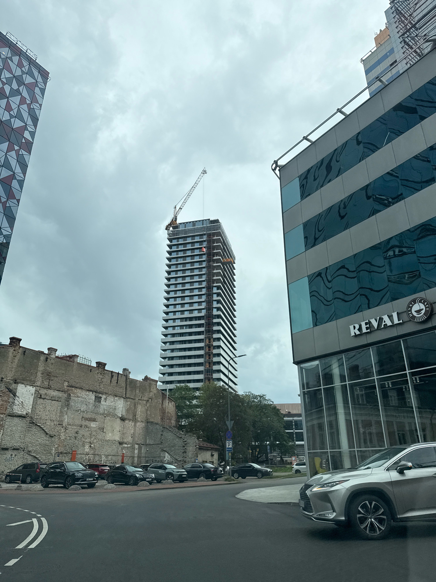 In Tallinn, Estonia, a street view shows a modern skyscraper under construction, topped with a crane. To its left, an older, partially demolished building with exposed brick walls stands. On the right, a contemporary building with reflective glass panels features a "REVAL CAFE" sign. Several cars are parked in front of the older structure, and a silver Lexus SUV is visible in the foreground on the street. The sky is overcast.