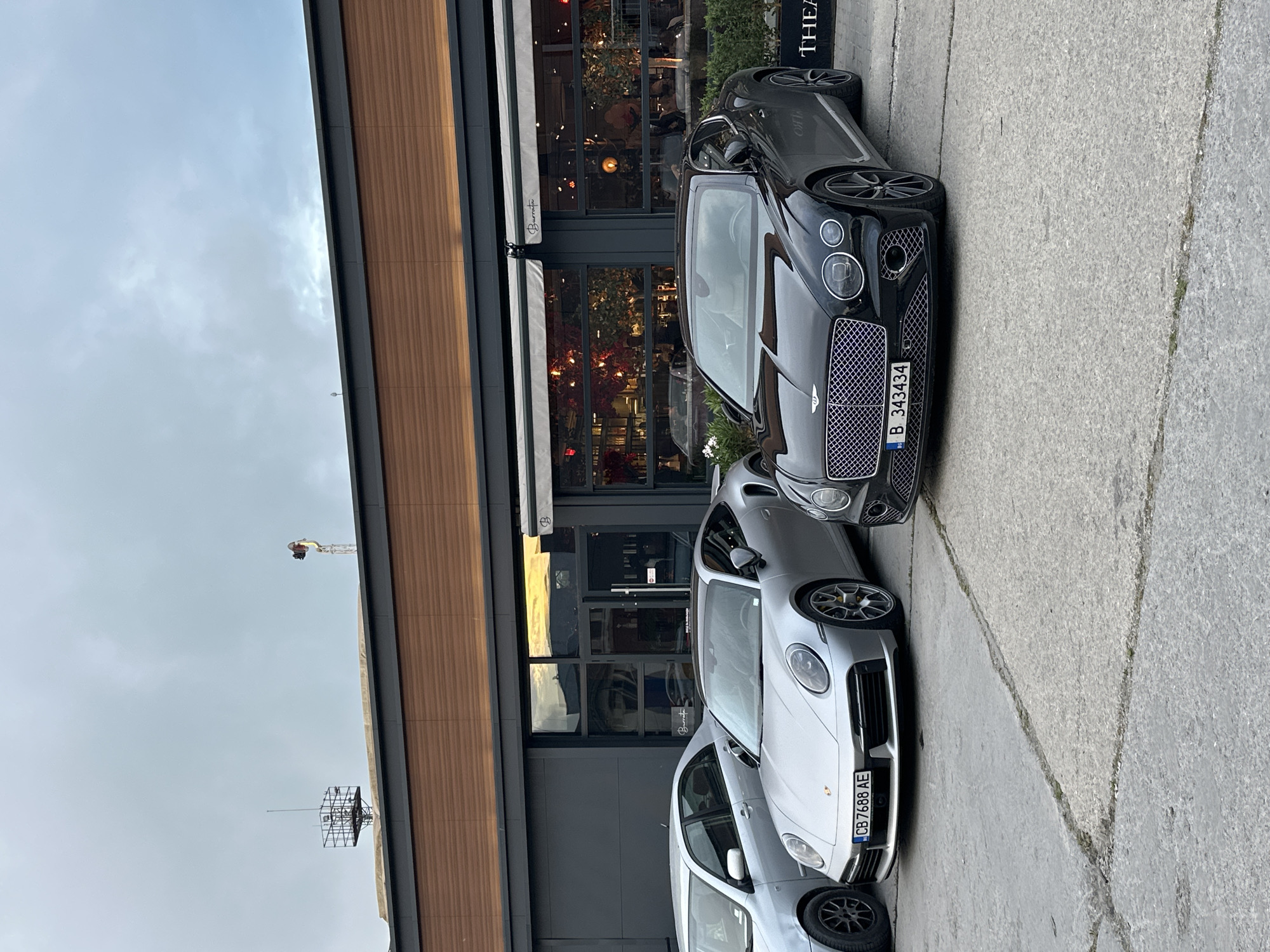 The image, correctly oriented, displays an urban street scene in Varna, Bulgaria, featuring several luxury cars parked alongside a modern building under an overcast sky. The lighting is soft and diffused, suggesting daytime without direct sunlight.
The main subjects are three high-end vehicles parked parallel to the building. Closest to the viewer is a silver Porsche 911, displaying the Bulgarian license plate "CB 7688 AE". Immediately behind it is a dark gray or black Bentley Continental GT, with the Bulgarian license plate "B 343434". Partially visible behind the Porsche is a lighter-colored sedan or SUV, and a dark car is partially seen behind the Bentley.
The vehicles are parked on a concrete pavement next to a contemporary building that appears to house commercial establishments. The building's lower section is characterized by large glass windows with dark frames, reflecting its surroundings. The upper facade features vertical wooden paneling. Through the windows, warm interior lighting is visible, suggesting a restaurant or cafe. An awning above one entrance prominently displays the name "Barriatou" in cursive script. A tall, black vertical sign to the right of the Bentley shows partial text, "THEA...", possibly indicating "THEATER".
In the distant background, against the cloudy sky, a construction crane and a metal structure, possibly an antenna or part of a building, are visible. The scene captures a static moment of urban luxury, with no immediate activity or people discernible.