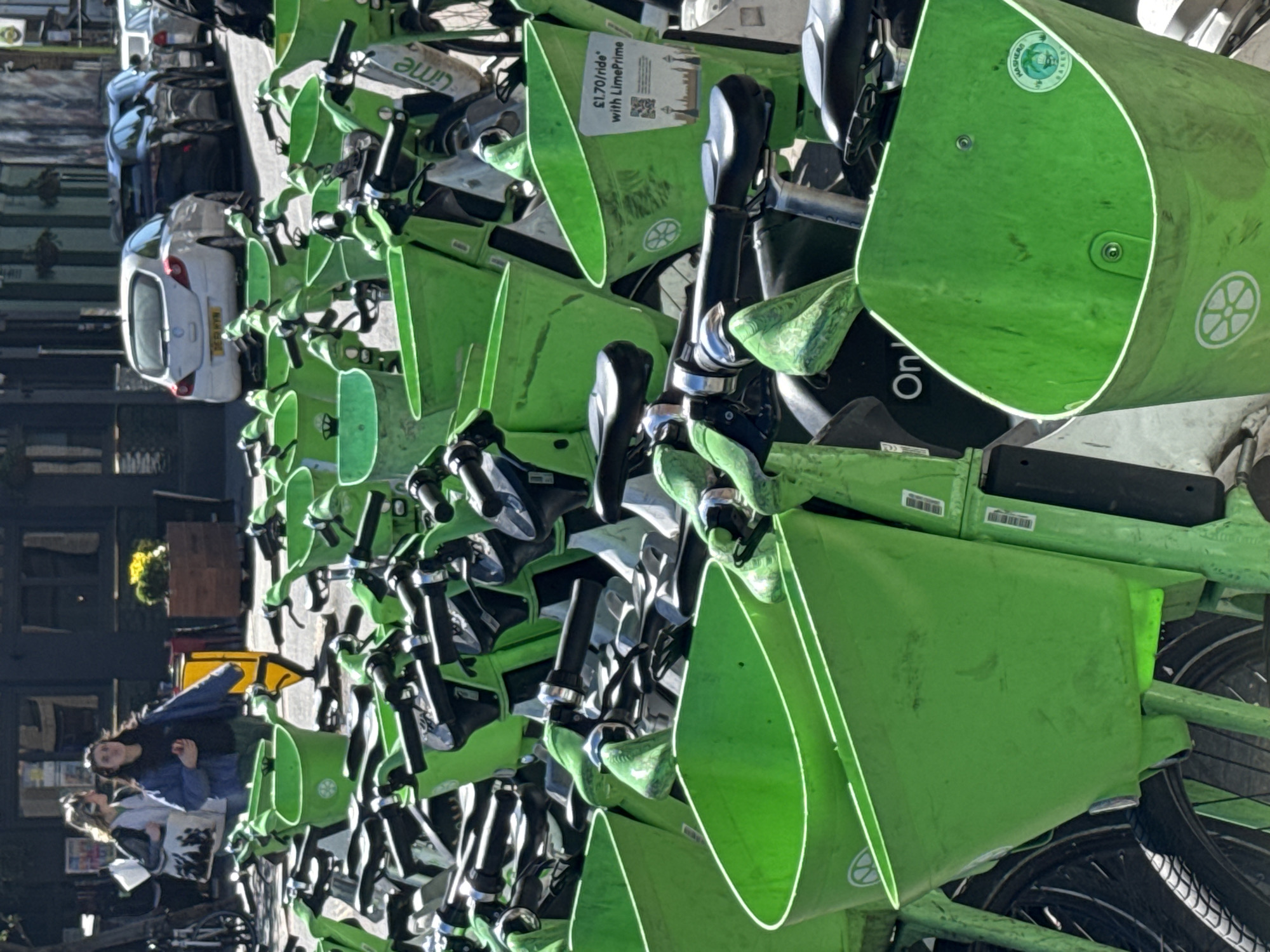 The image, set in an urban environment within the United Kingdom (consistent with the provided London context), captures a dense arrangement of bright green Lime rental bicycles and electric scooters parked on a paved surface, likely a sidewalk or dedicated bay. The scene is illuminated by natural daylight, suggesting a clear and sunny day.
The foreground and mid-ground are dominated by these numerous green vehicles, many equipped with front baskets or platforms. Prominently displayed on one basket is a sign detailing pricing: "£1.70/ride with LimePrime," accompanied by a QR code for rental access. Another green basket features a circular sticker with the text "MAIDENHEAD BIKES," indicating a specific operational area or program within the UK. The "Lime" brand logo, resembling a wheel, is visible on several baskets. Barcodes and serial numbers are also visible on the frames of some vehicles.
In the background, a white car with a UK license plate, "DE53 HTN," is parked on the street, confirming the UK setting. Other vehicles and building facades are partially visible further down the street. On the left side of the image, two individuals, likely women, are standing near the bikes. One wears a blue jacket, and the other a patterned jacket. They appear to be casually observing or waiting, with no strong emotions evident. The overall impression is a busy, typical daytime street scene featuring a popular shared mobility service.