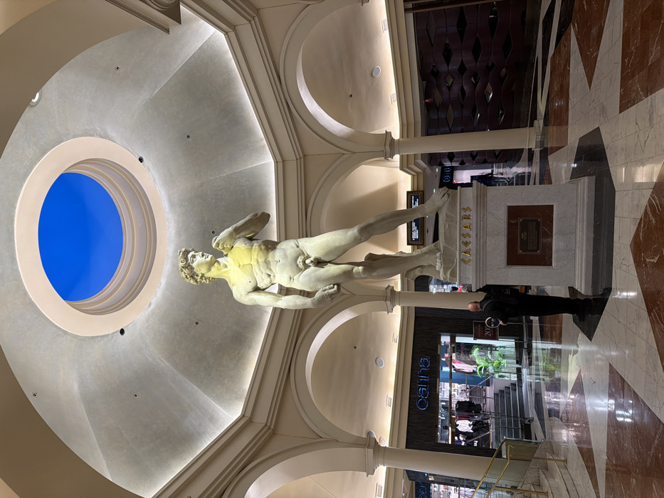An interior view features a marble statue of David under a domed ceiling with a round opening showcasing a bright blue sky. The statue stands on a pedestal labeled "CAESARS." A man in a black suit stands near the base. The architecture includes arches and decorative pillars. The floor is a patterned marble. A clothing store is visible in the background.